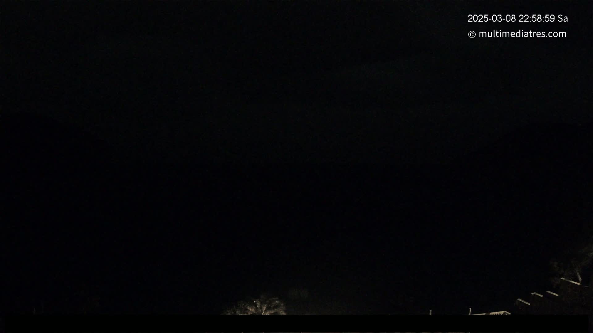 A nighttime, dark view shows a few dimly lit structures at the bottom of the frame, under a completely black sky.