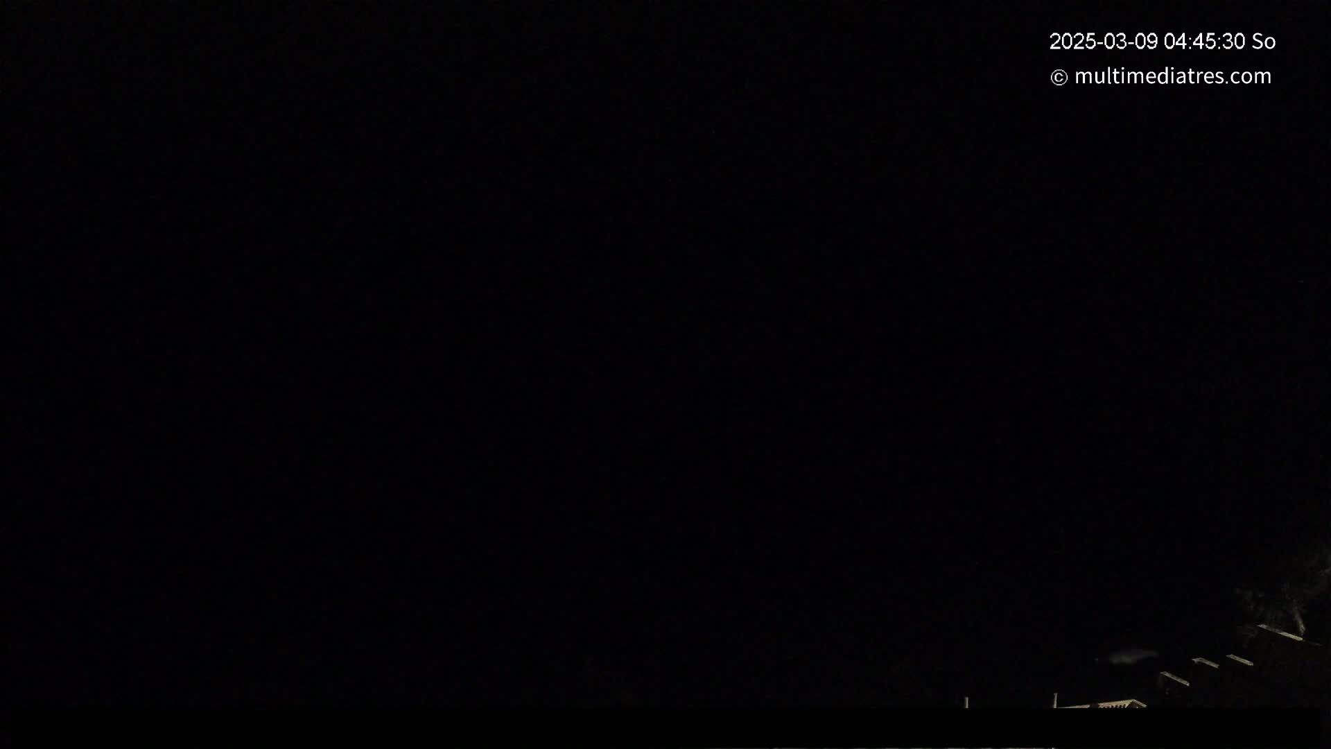 A nighttime image shows a dark, mostly clear sky with the silhouettes of some rooftops and trees at the bottom of the frame.