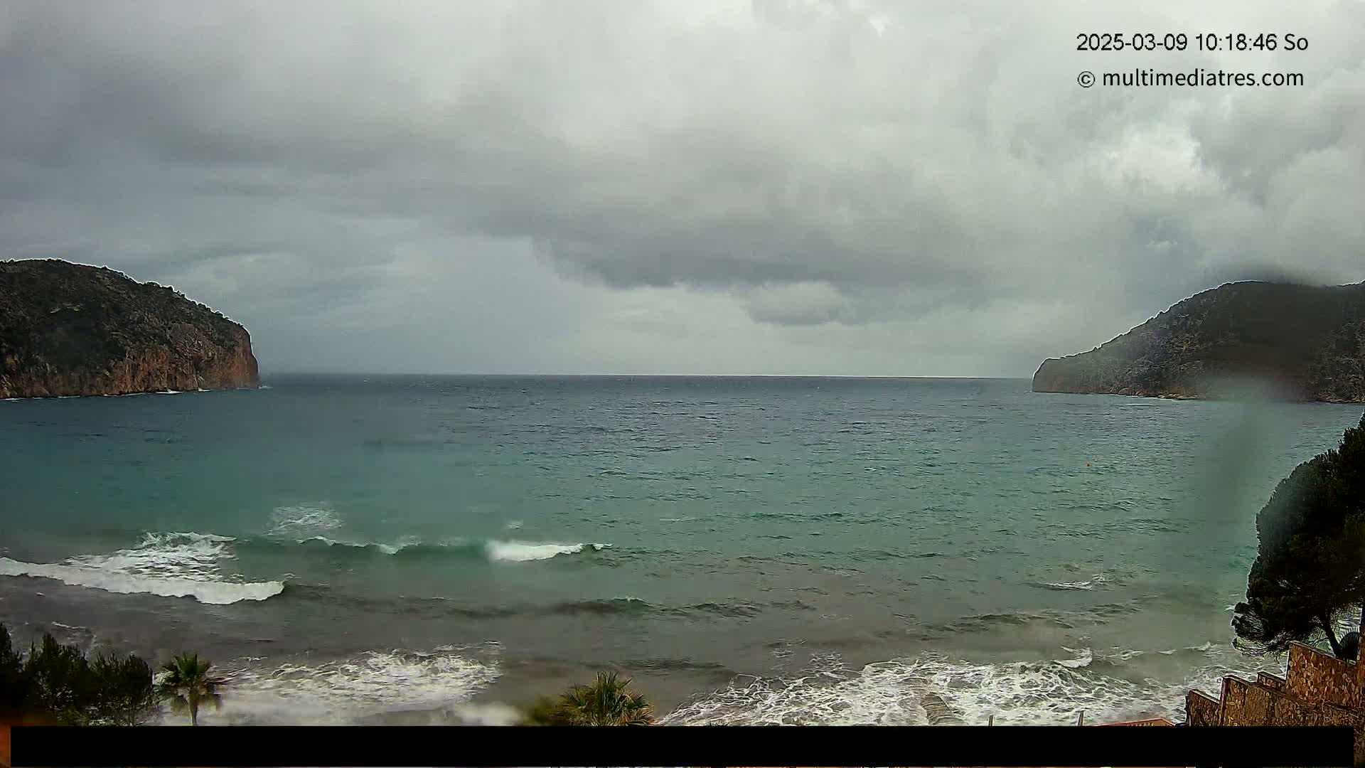 A body of water with small waves laps against a shore under an overcast sky, framed by rocky hills on either side.