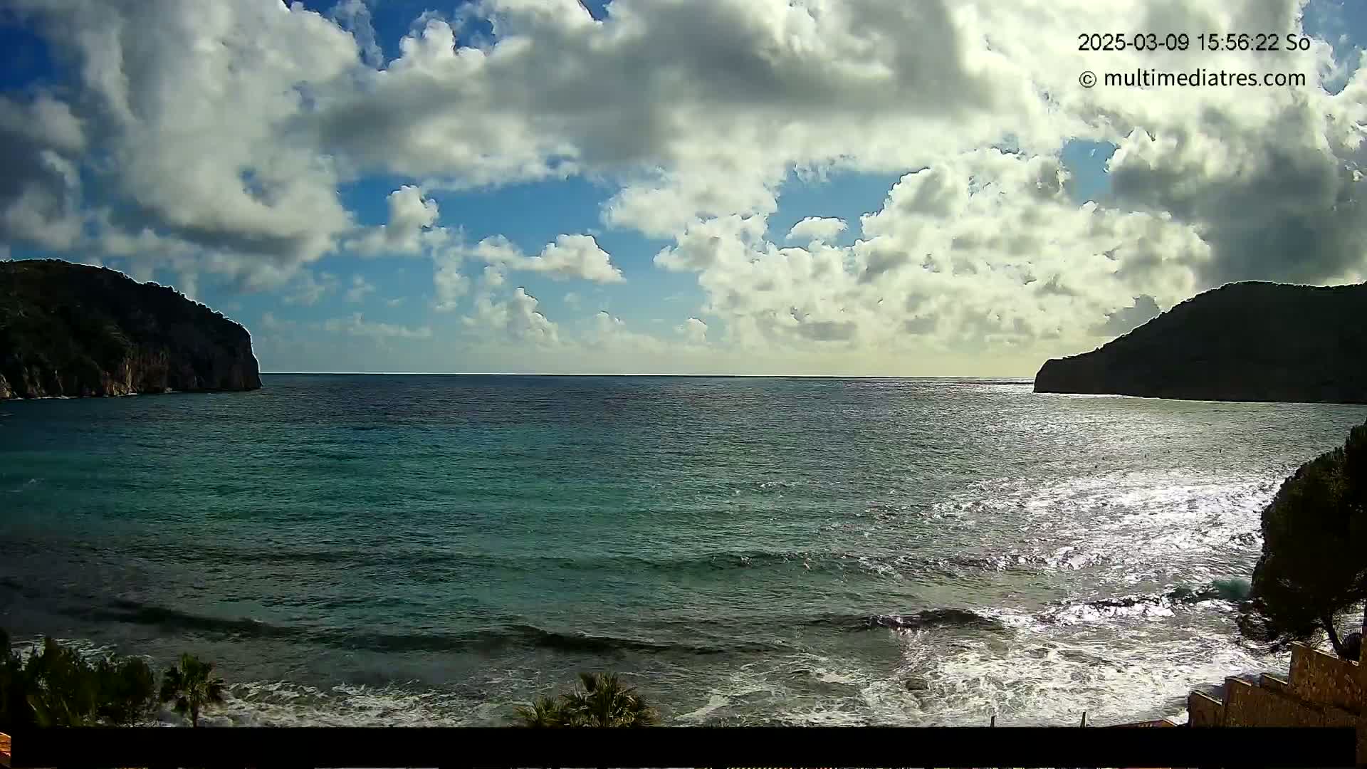 A partly cloudy day shows a tranquil ocean scene with dark green water lapping gently on a shore between two dark, rocky hills.