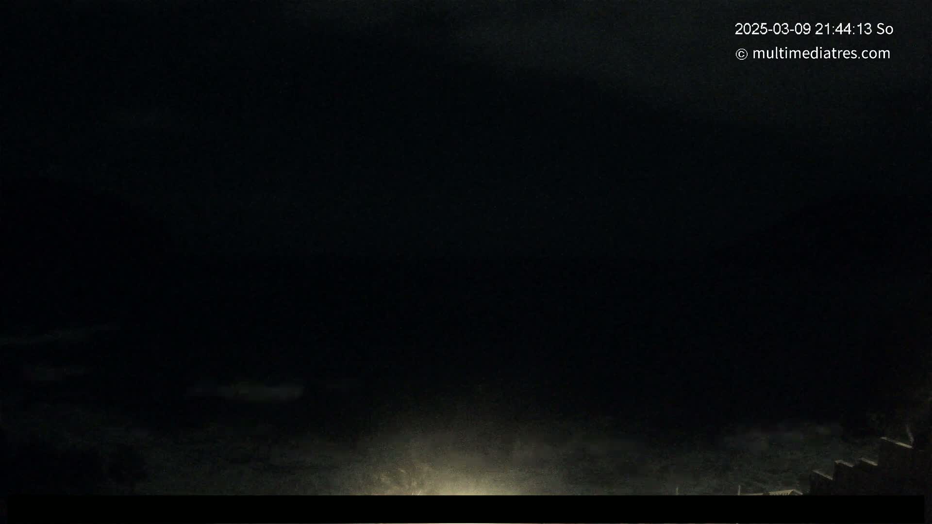 A dark, nighttime scene shows a body of water and some land in the lower portion of the frame, under an overcast sky.