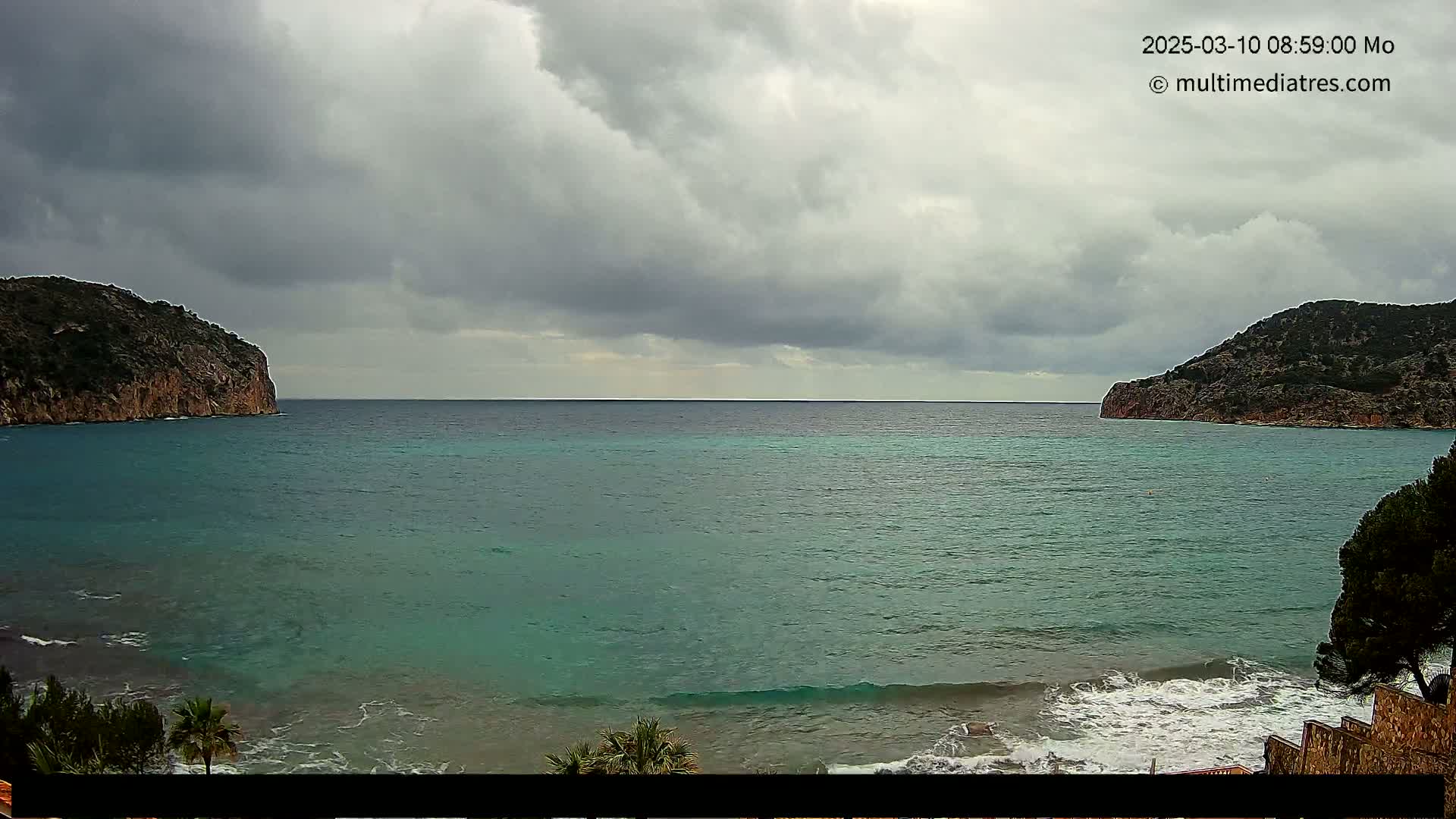A teal sea with small waves laps a beach between two rocky hills under a cloudy sky.