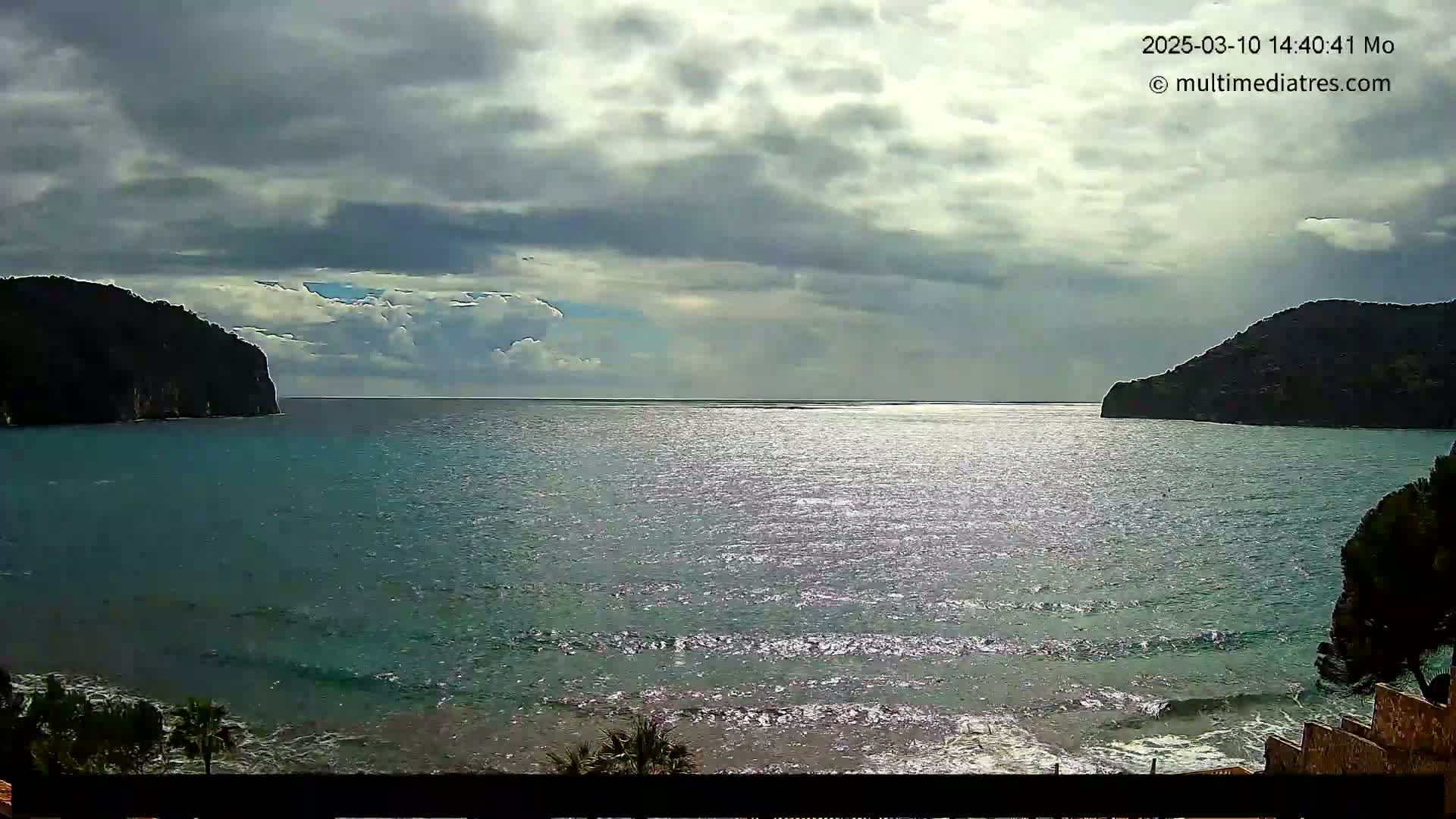A partly cloudy day shows a calm sea between two dark, rocky hillsides, with small waves lapping at a sandy shore.