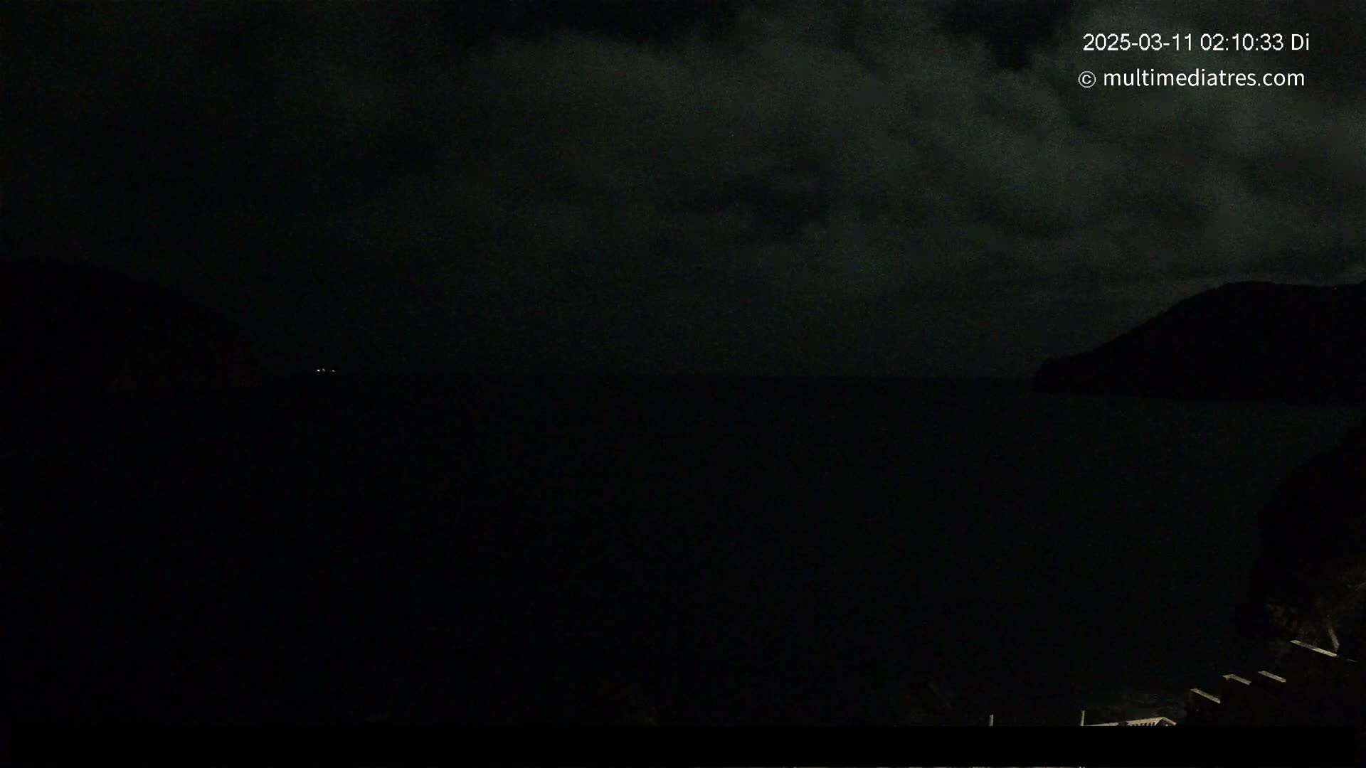 A dark, nighttime coastal scene shows a body of water between two hills under a cloudy sky.