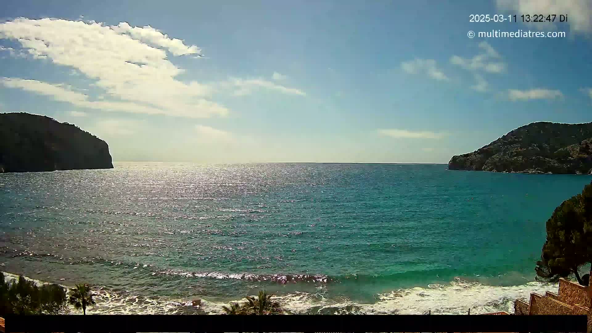 A partly cloudy sunny day shows a picturesque coastal scene of a teal ocean with small waves lapping a sandy shore, framed by dark, rocky hillsides.