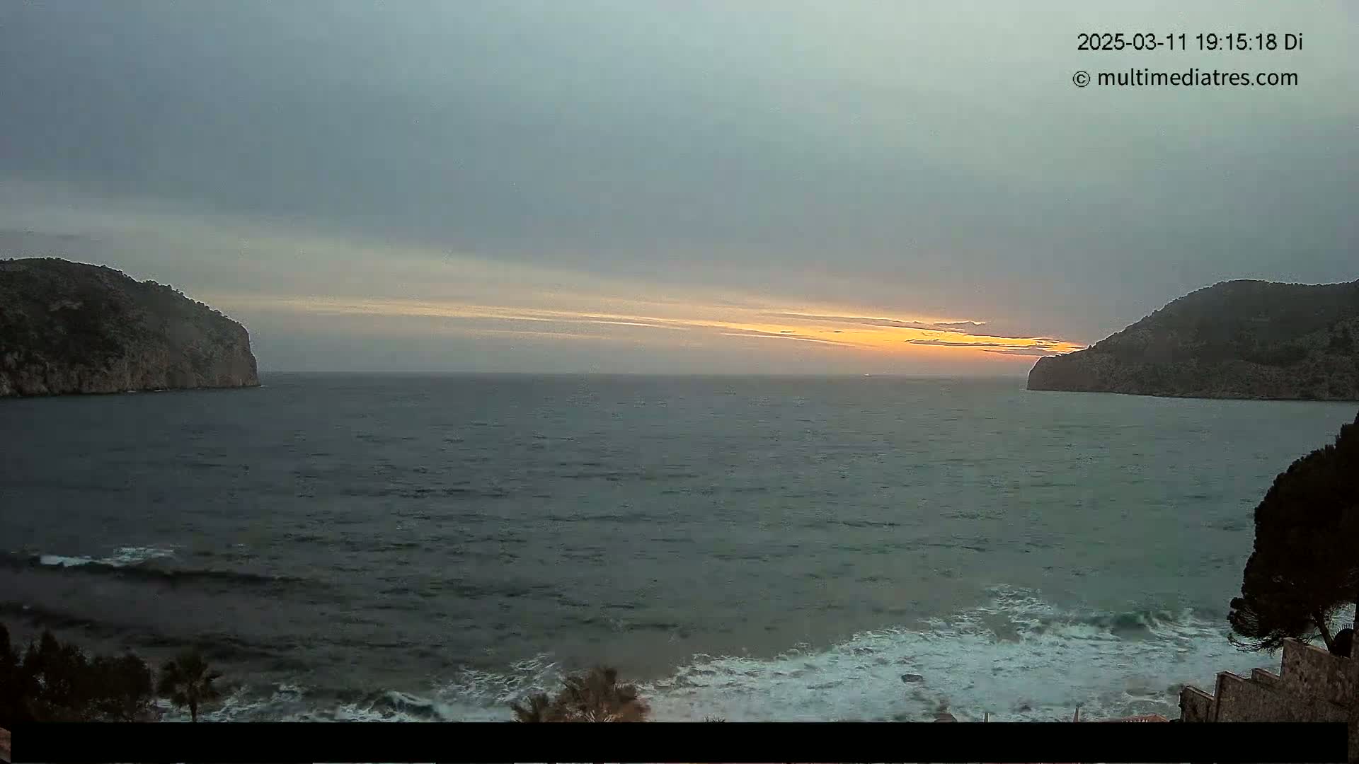 A partly cloudy sunset over a dark, wavy ocean, with landmasses on either side of the bay.