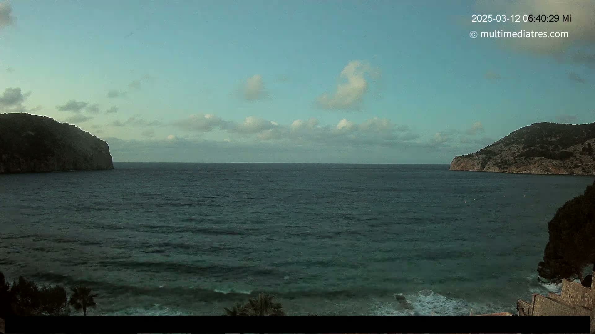 A calm sea is seen between two dark, rocky headlands under a mostly clear sky with a few scattered clouds.