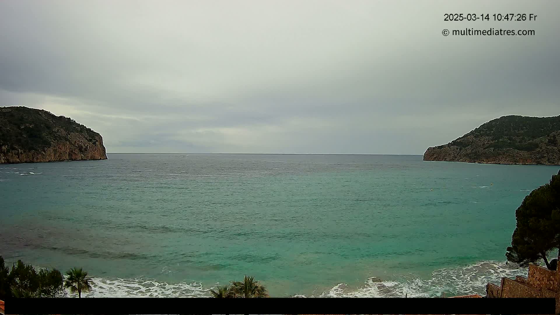 A calm, teal sea is nestled between two rocky hills under an overcast sky, with small waves breaking on a sandy shore.