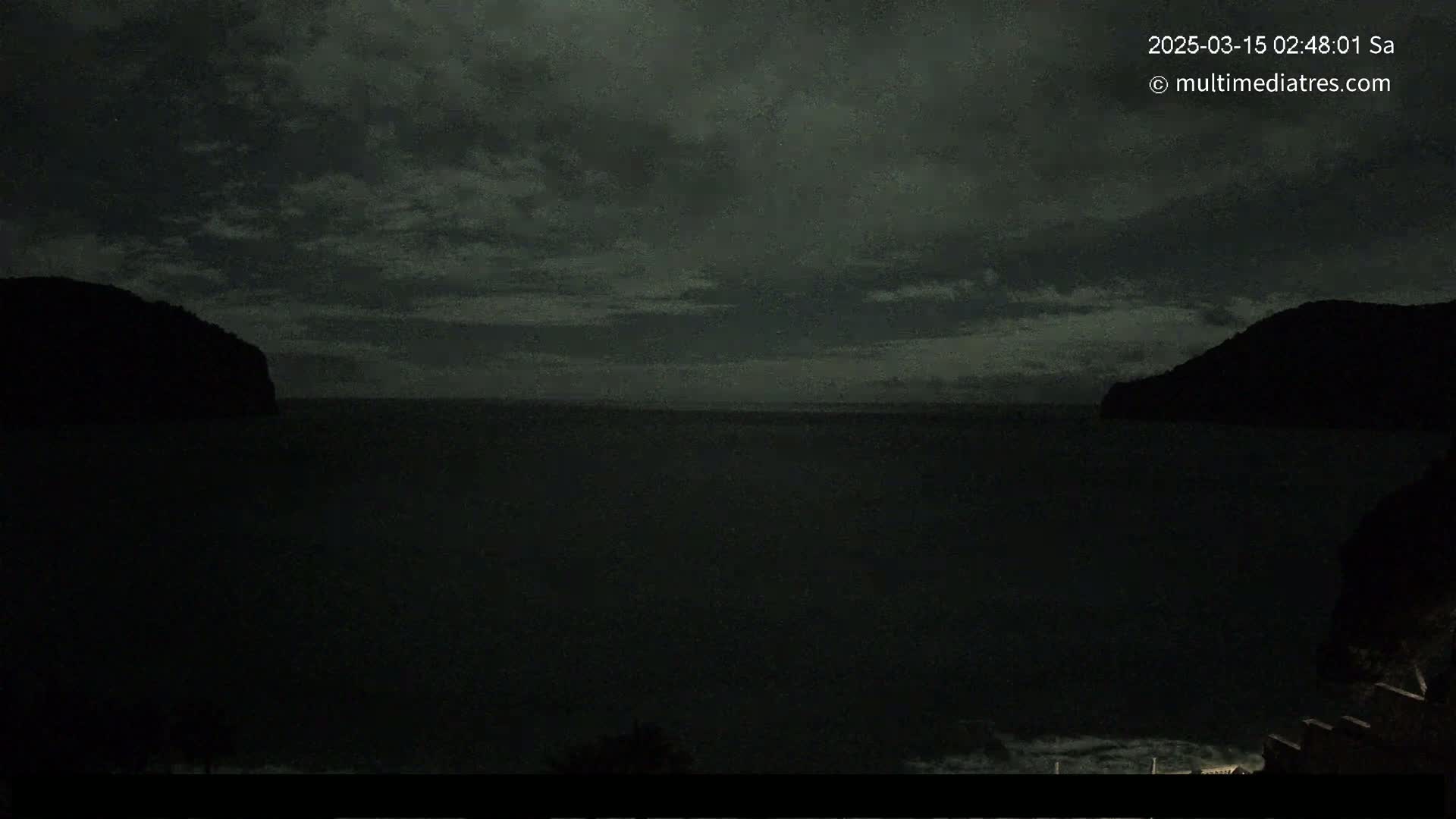 A dark, nighttime view of a calm body of water between two silhouetted hills under a cloudy sky.