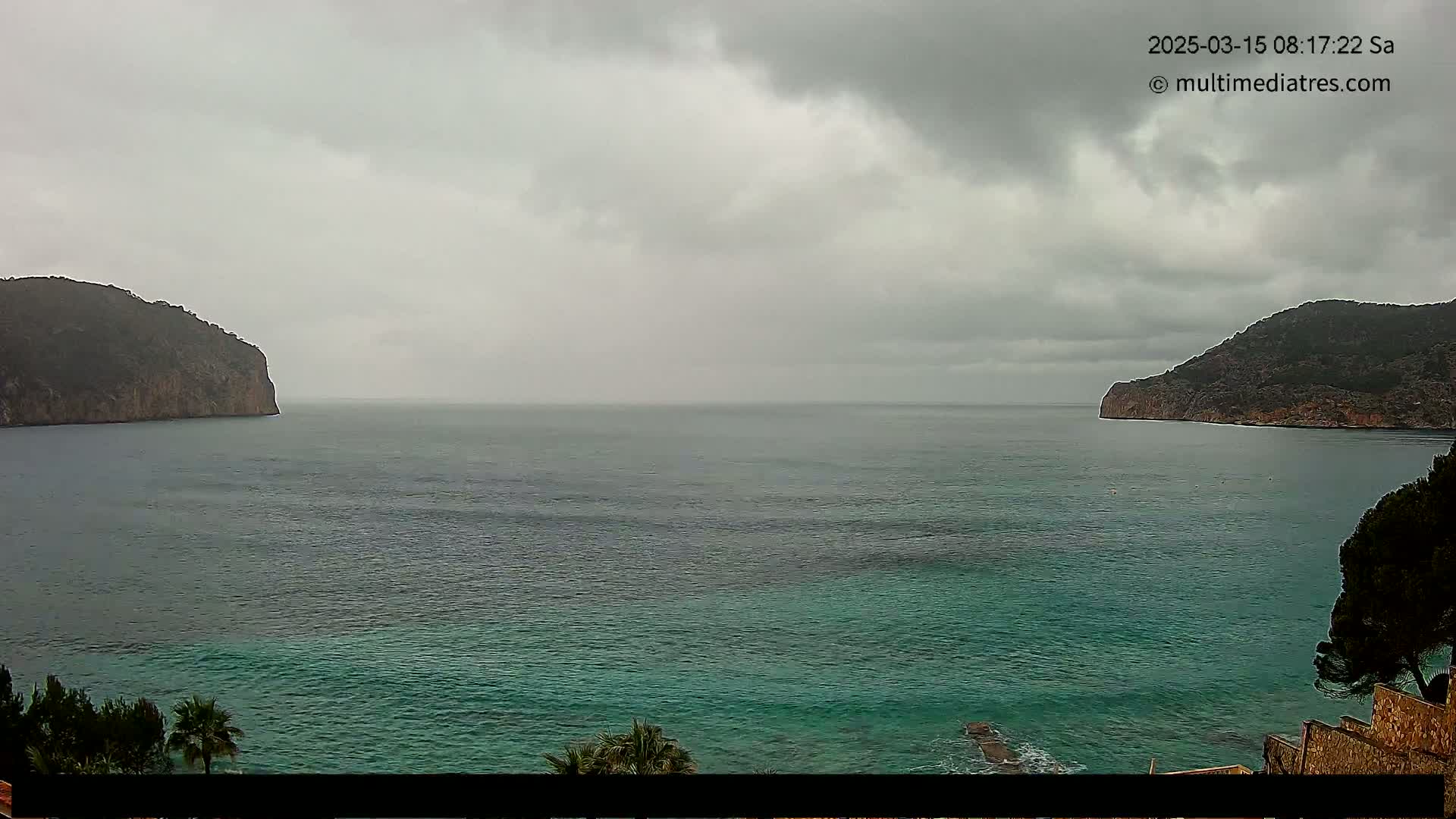 A calm, overcast day shows a view of a teal sea between two dark, rocky cliffs, with a bit of greenery visible in the foreground.