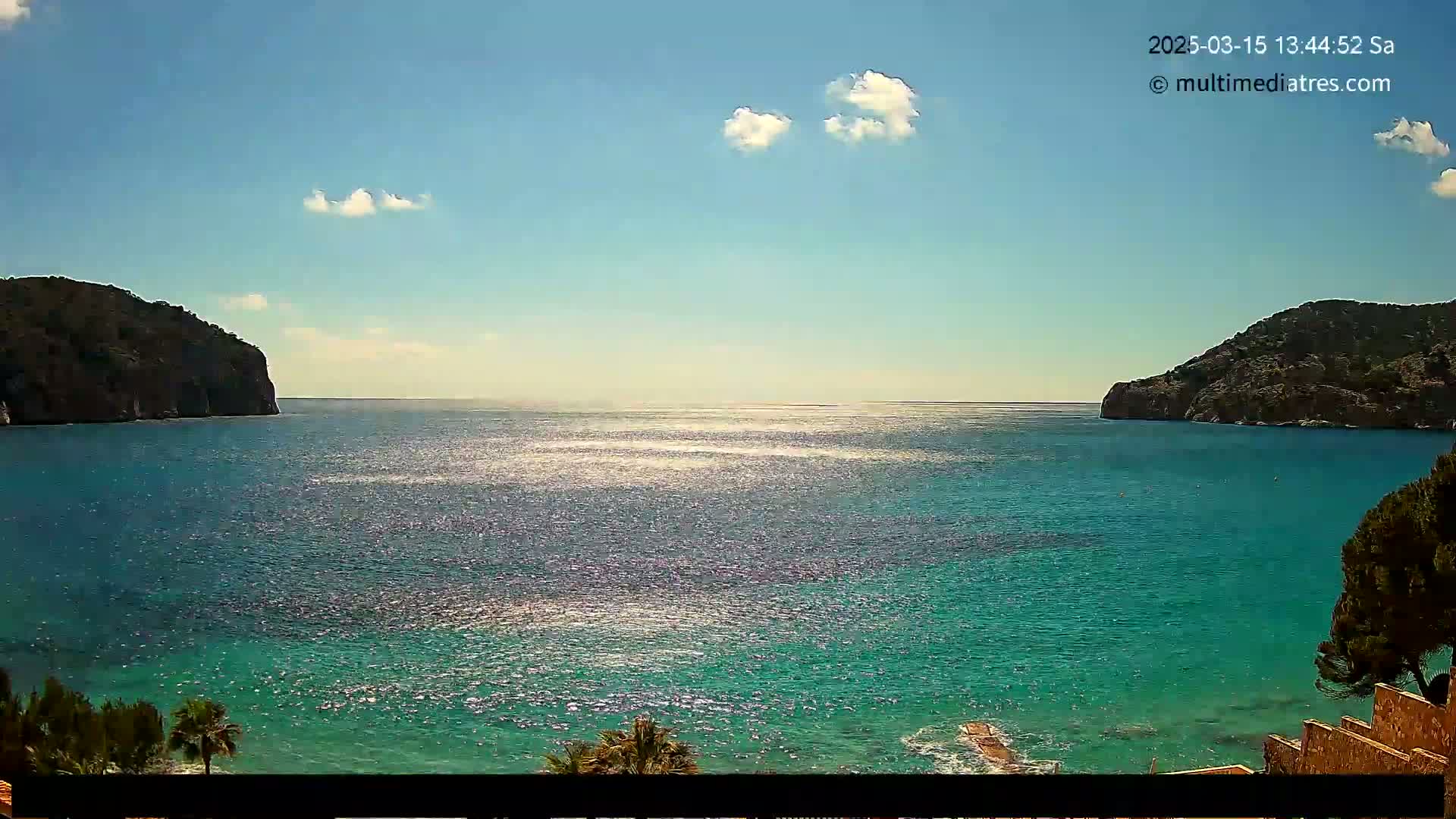 A sunny day reveals a sparkling turquoise ocean cove nestled between two dark, rocky cliffs, with a few fluffy clouds in the sky.