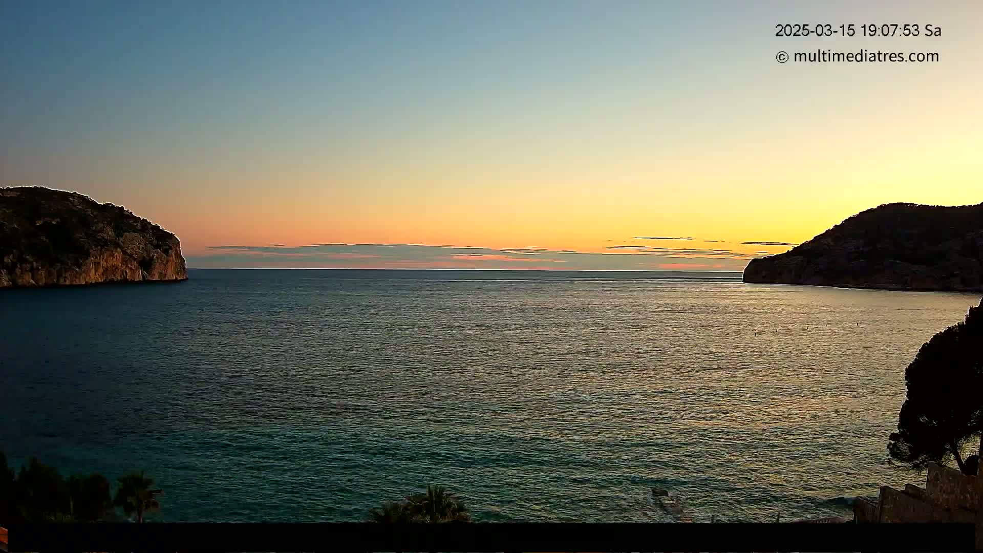 Two dark, rocky hillsides frame a calm sea at sunset under a clear, pale orange and blue sky.
