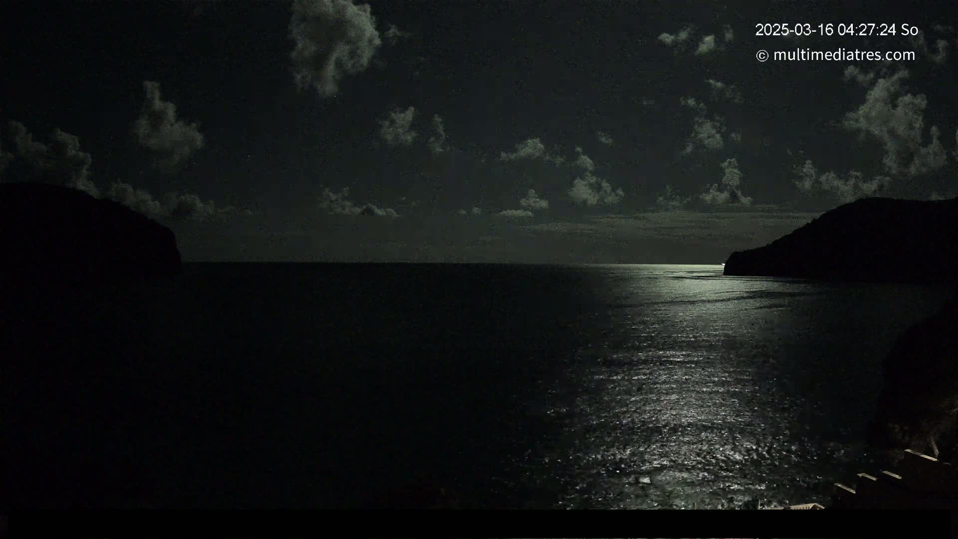 A moonlit night scene shows a dark body of water between two silhouetted hills under a partly cloudy sky.