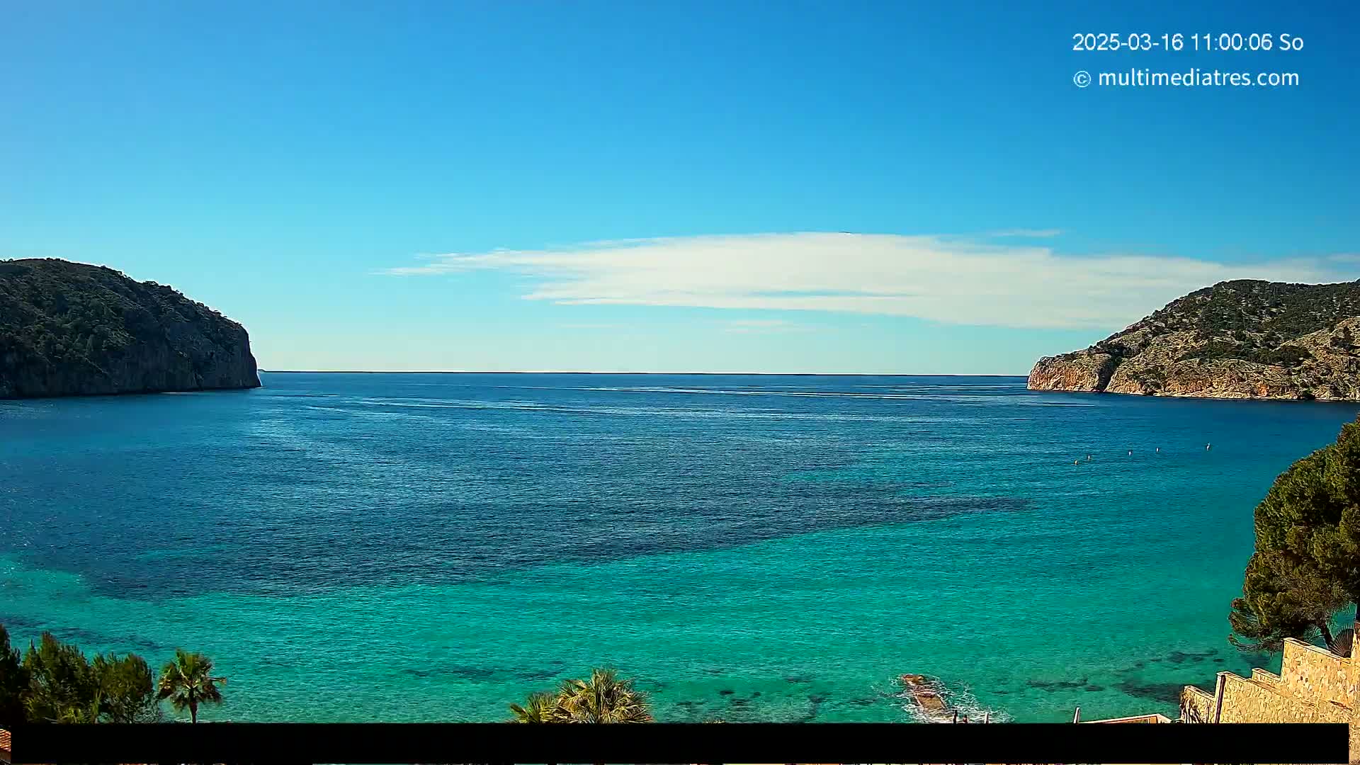 A calm, bright blue sea with varying shades of turquoise near the shore, nestled between two rocky hills under a mostly clear sky.