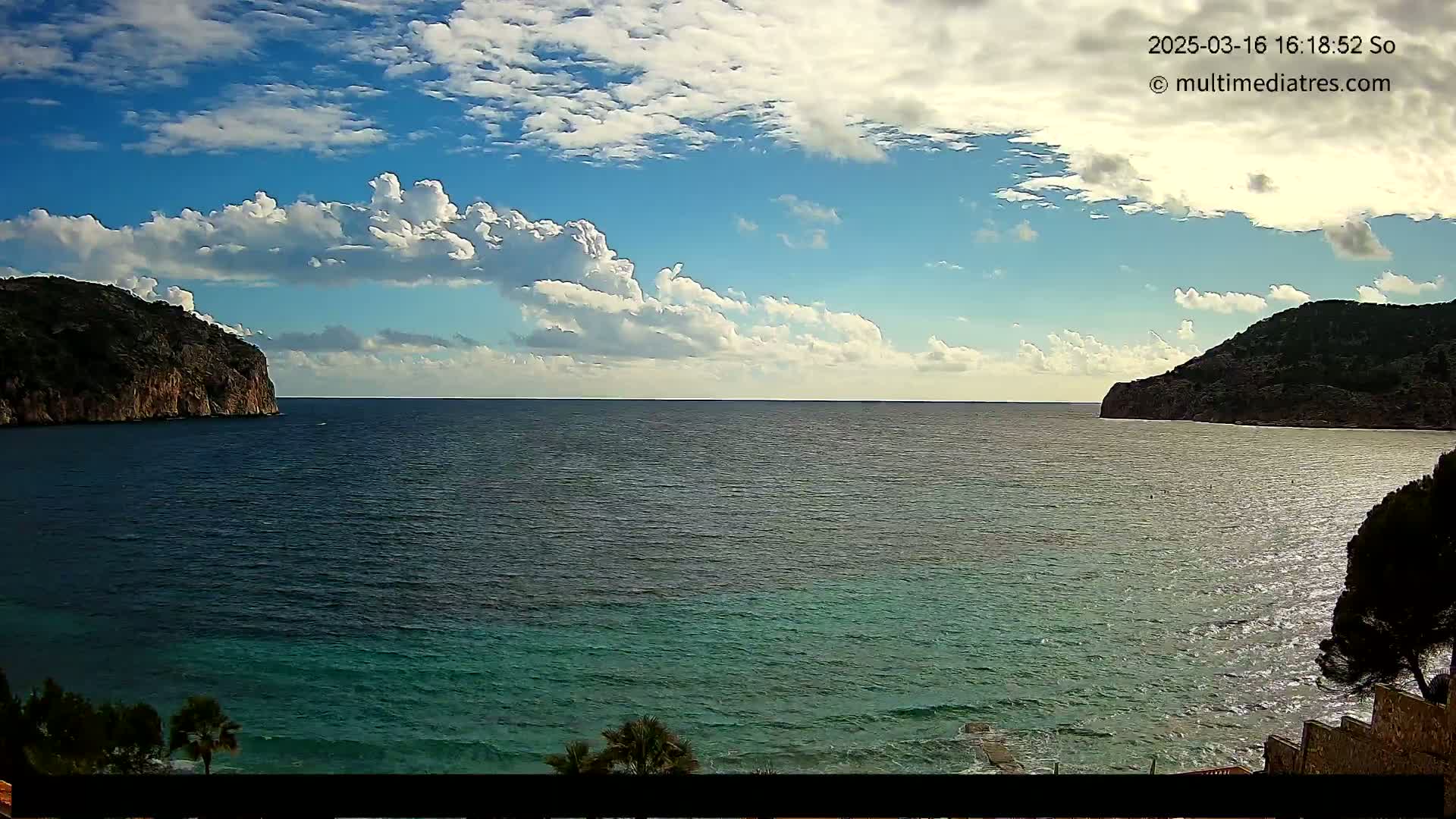 A partly cloudy sky overlooks a calm sea between two dark, rocky hills.