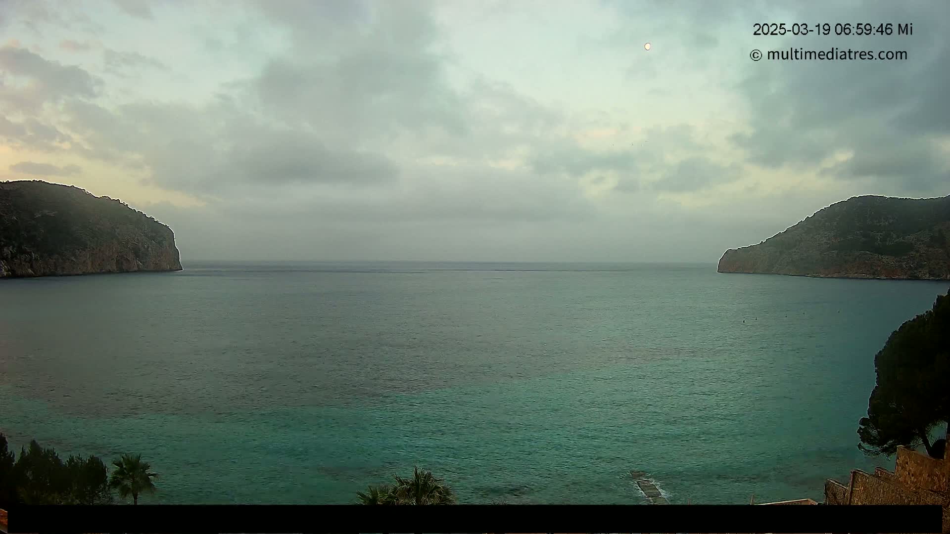 A calm, overcast day shows a teal-colored bay bordered by two dark, rocky headlands.