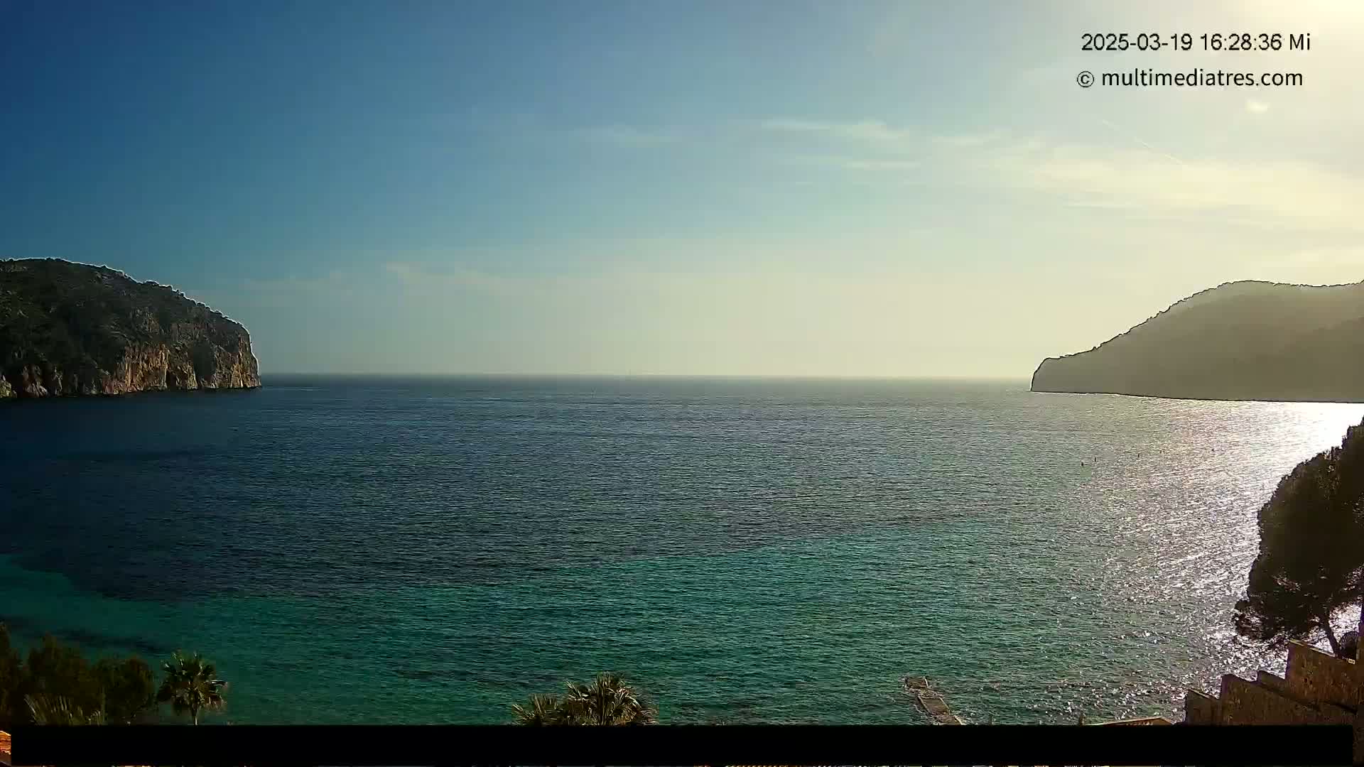 A calm sea with clear, teal-green water is seen between two dark, rocky hills under a mostly sunny sky.