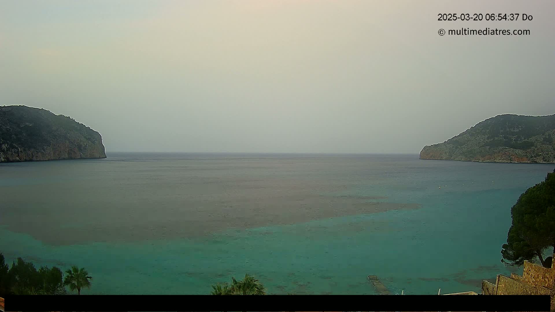 A hazy, calm bay is nestled between two dark, rocky hills, with teal-colored water visible near the shore.