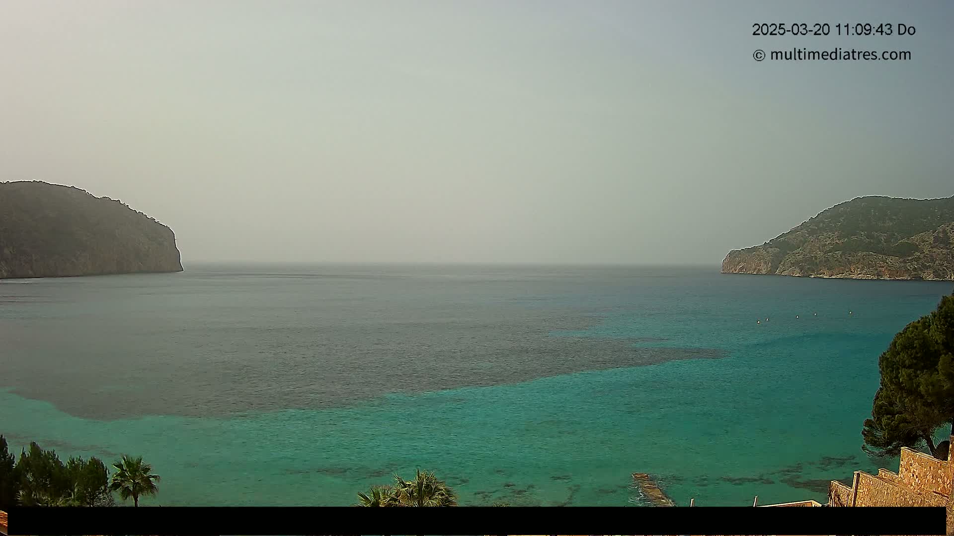 A hazy view of a calm, teal-colored bay nestled between two rocky hills.