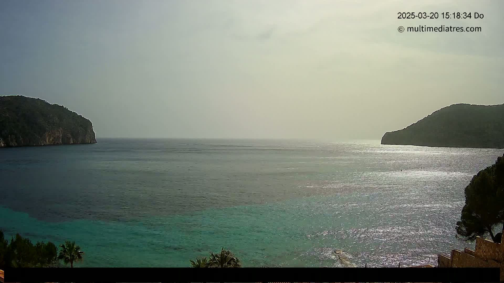A hazy, sunny day reveals a calm bay of teal water between two dark green, rocky hillsides.