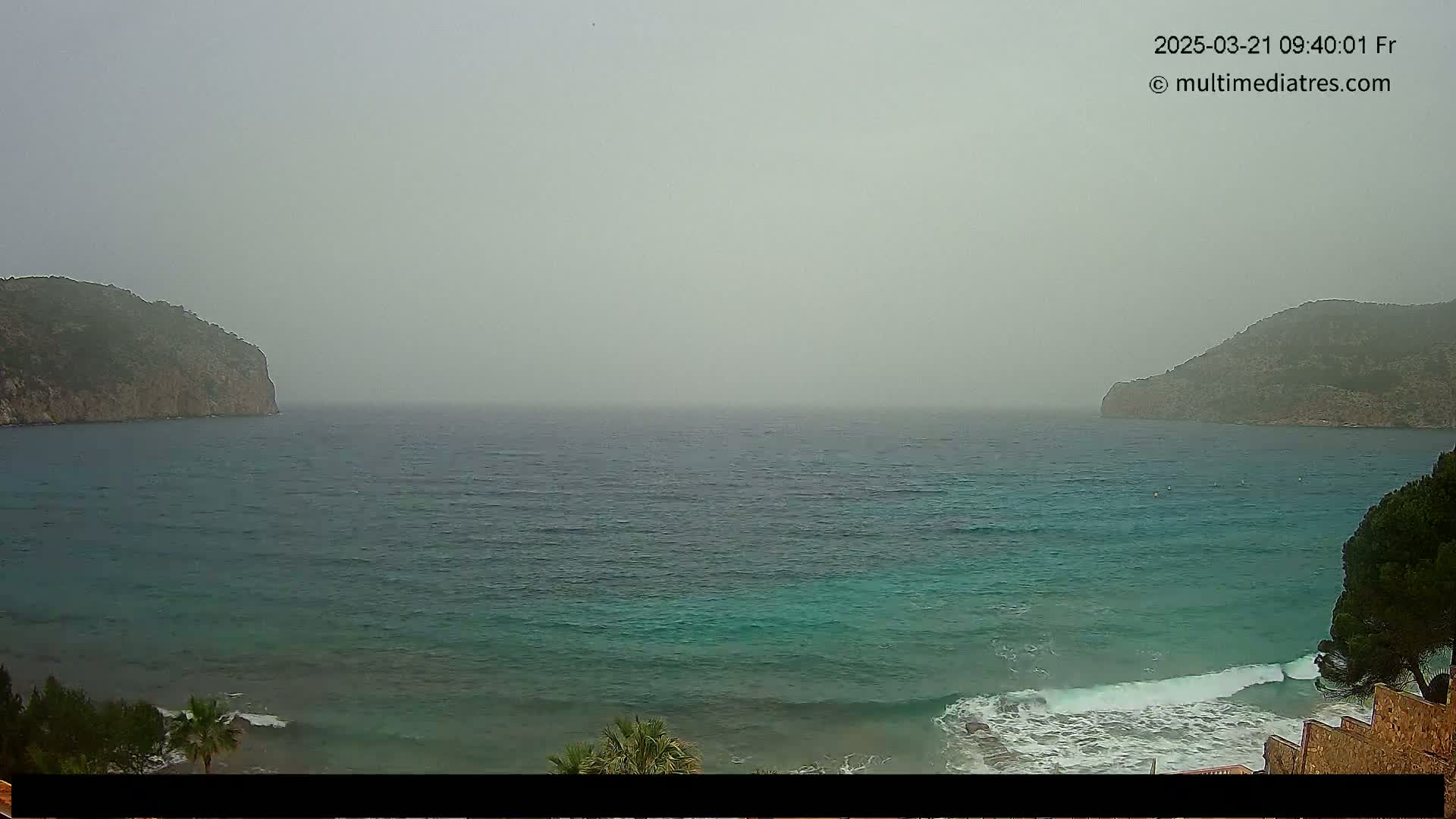 A hazy, overcast day reveals a teal sea lapping at a rocky shore between two low, dark-green hills.