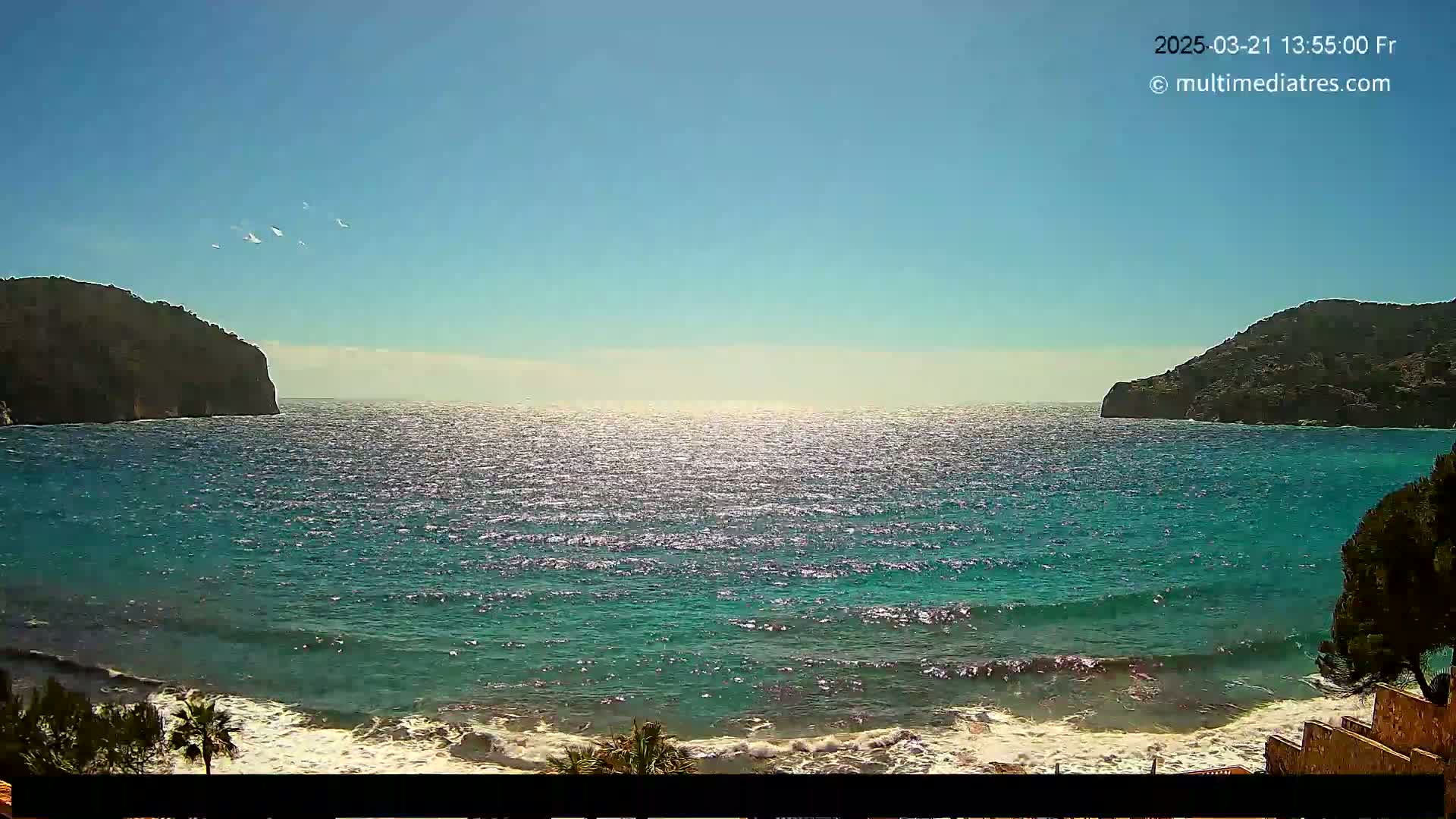 A sunny day shows a tranquil teal ocean with gentle waves lapping a shore between two dark, rocky headlands.