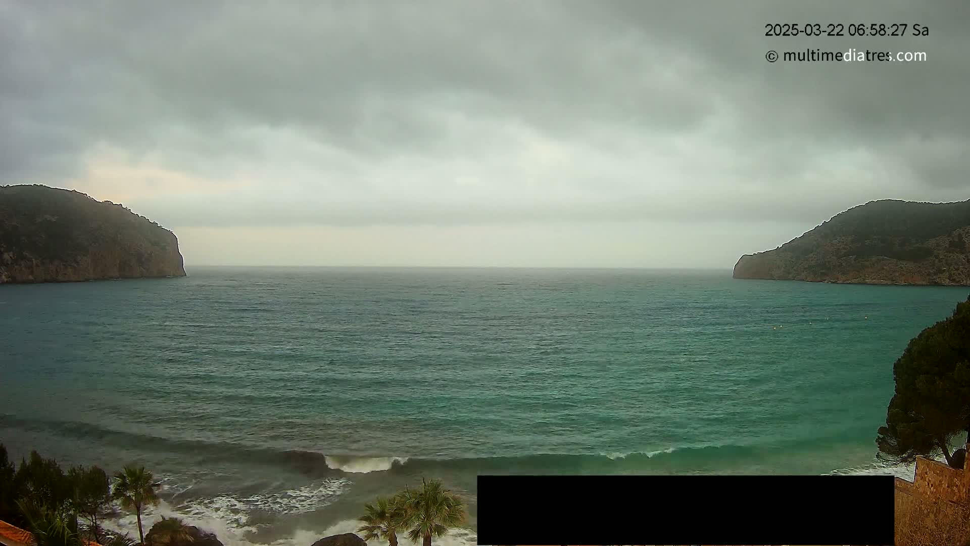 A body of teal water with small waves laps against a rocky shore under an overcast sky, framed by two dark hills.