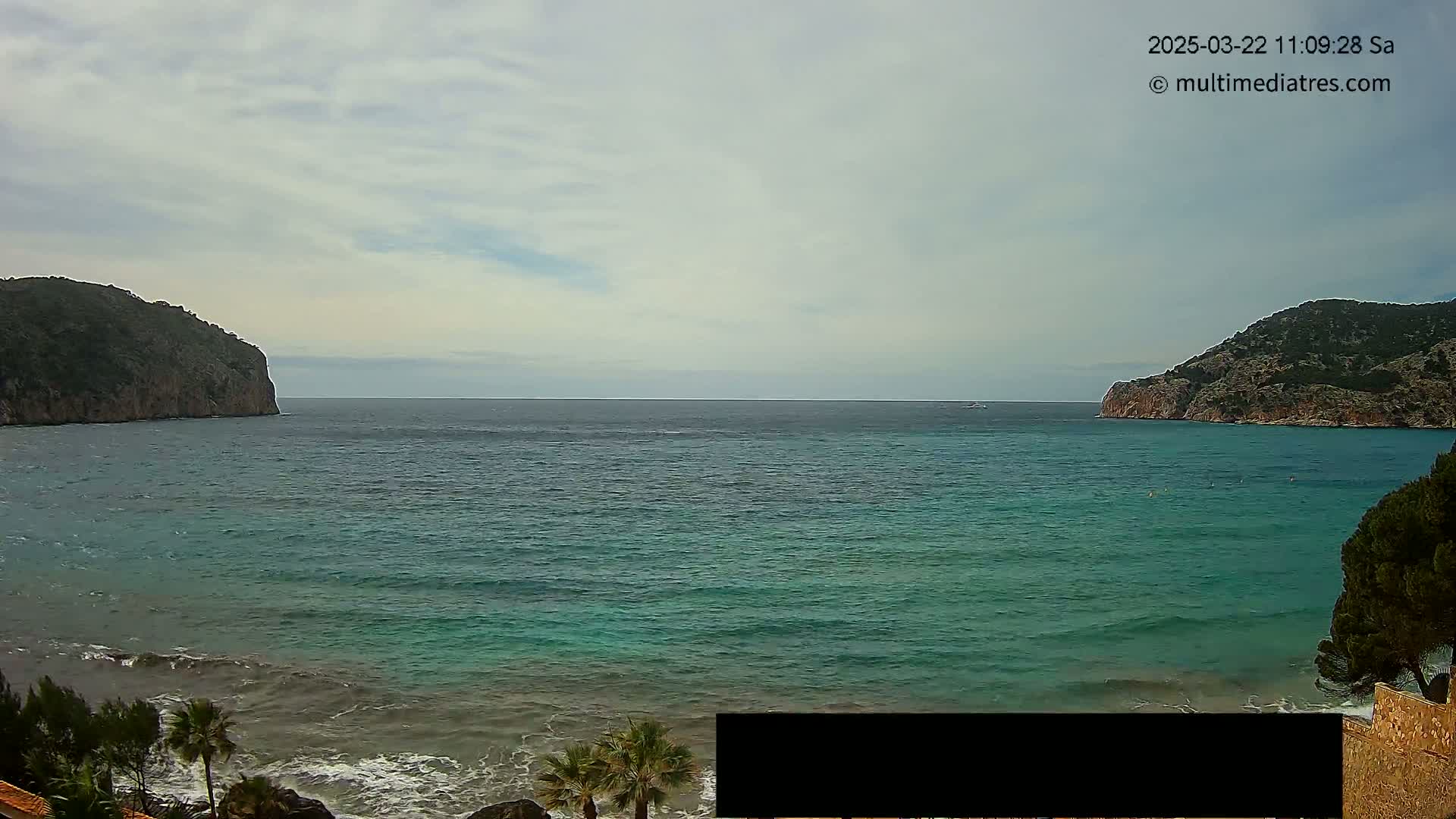 A calm, partly cloudy day reveals a tranquil bay of teal water nestled between two rocky cliffs, with small waves gently lapping at the shore.