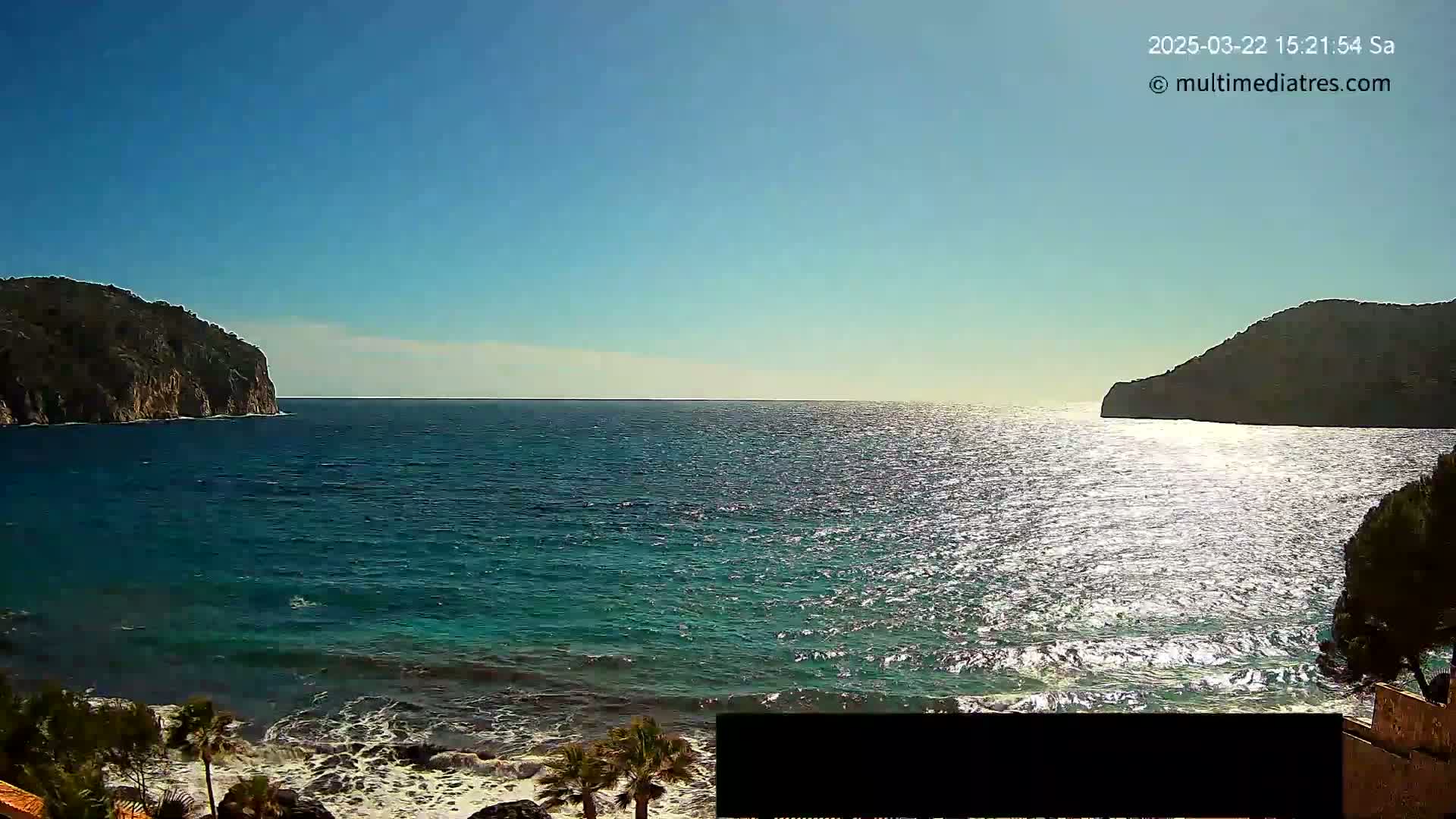Under a clear blue sky, a tranquil ocean scene unfolds, featuring dark, rocky cliffs on either side of a sparkling, wave-kissed body of water.