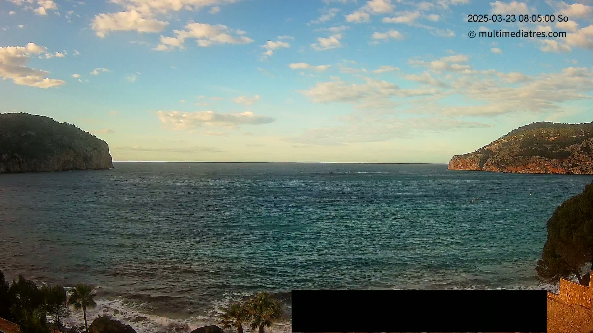 A calm sea with partly cloudy skies is seen between two rocky, tree-covered headlands.