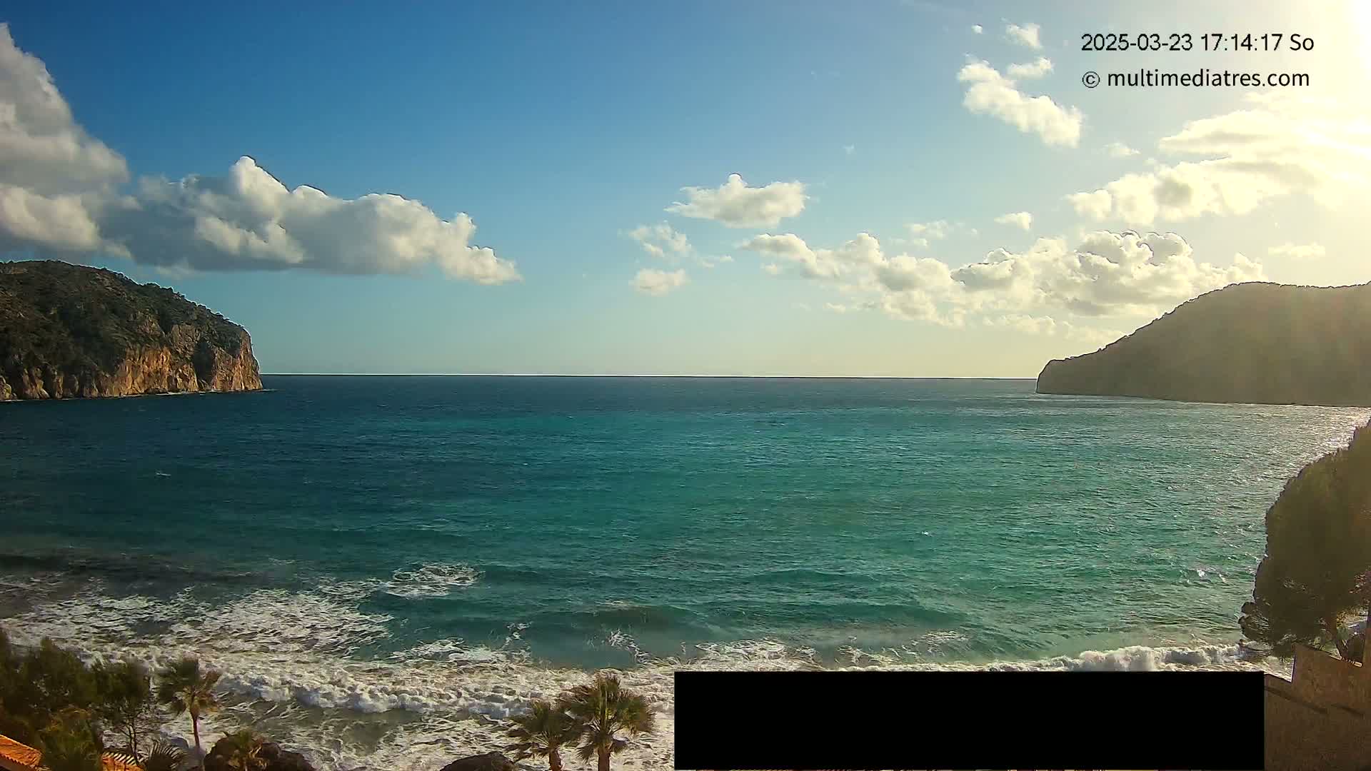 A partly cloudy day shows a view of a teal ocean with waves breaking on a beach, framed by dark cliffs on either side.