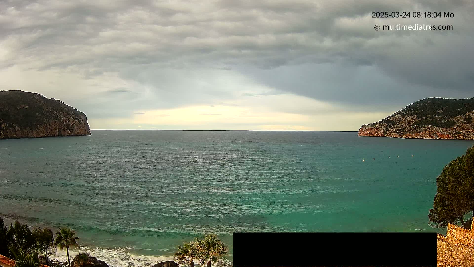 A calm, teal sea is nestled between two rocky hills under a cloudy sky.