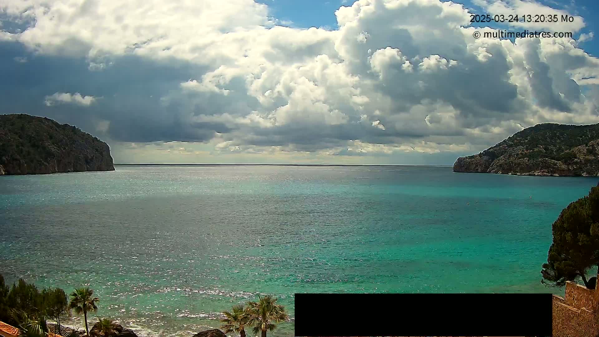 A partly cloudy sky overlooks a calm, teal-colored bay nestled between two rocky hillsides.