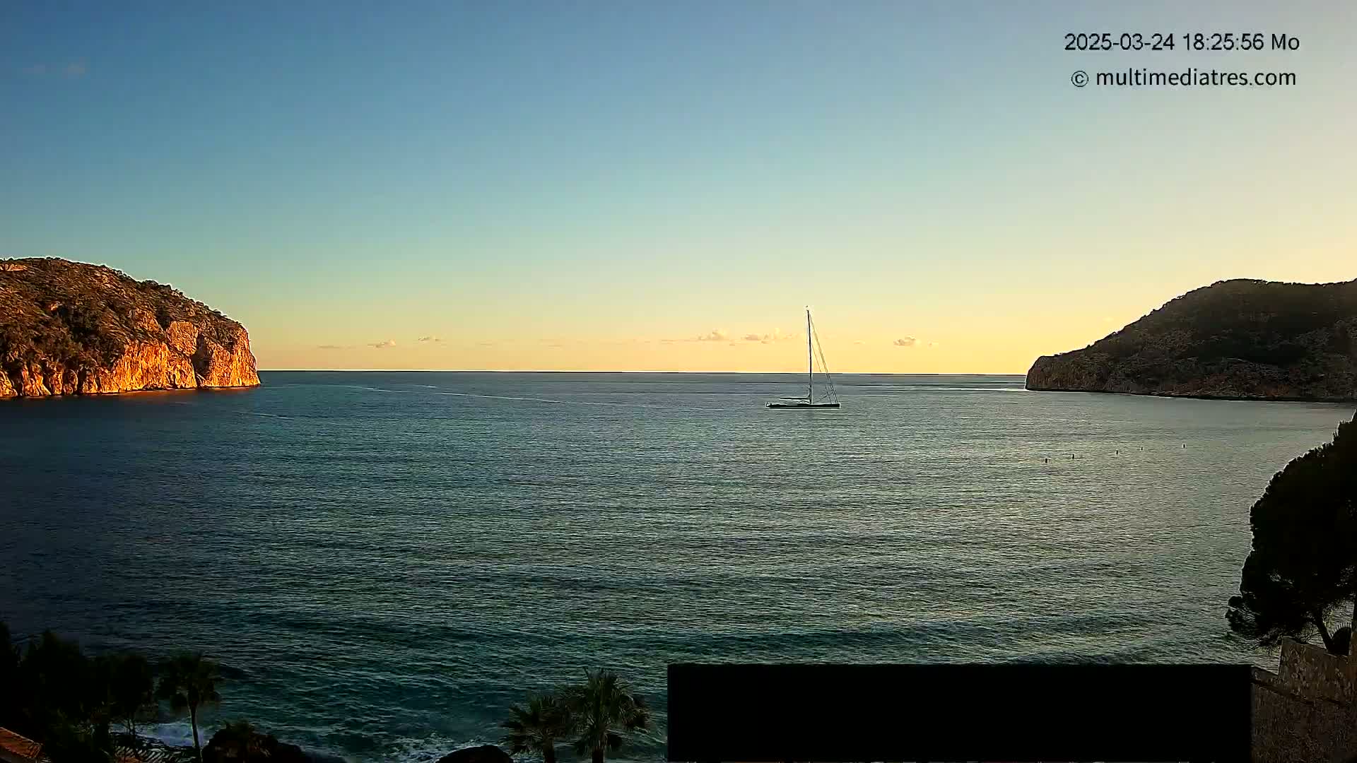A sailboat is visible on a calm sea at sunset, between two dark, rocky headlands.