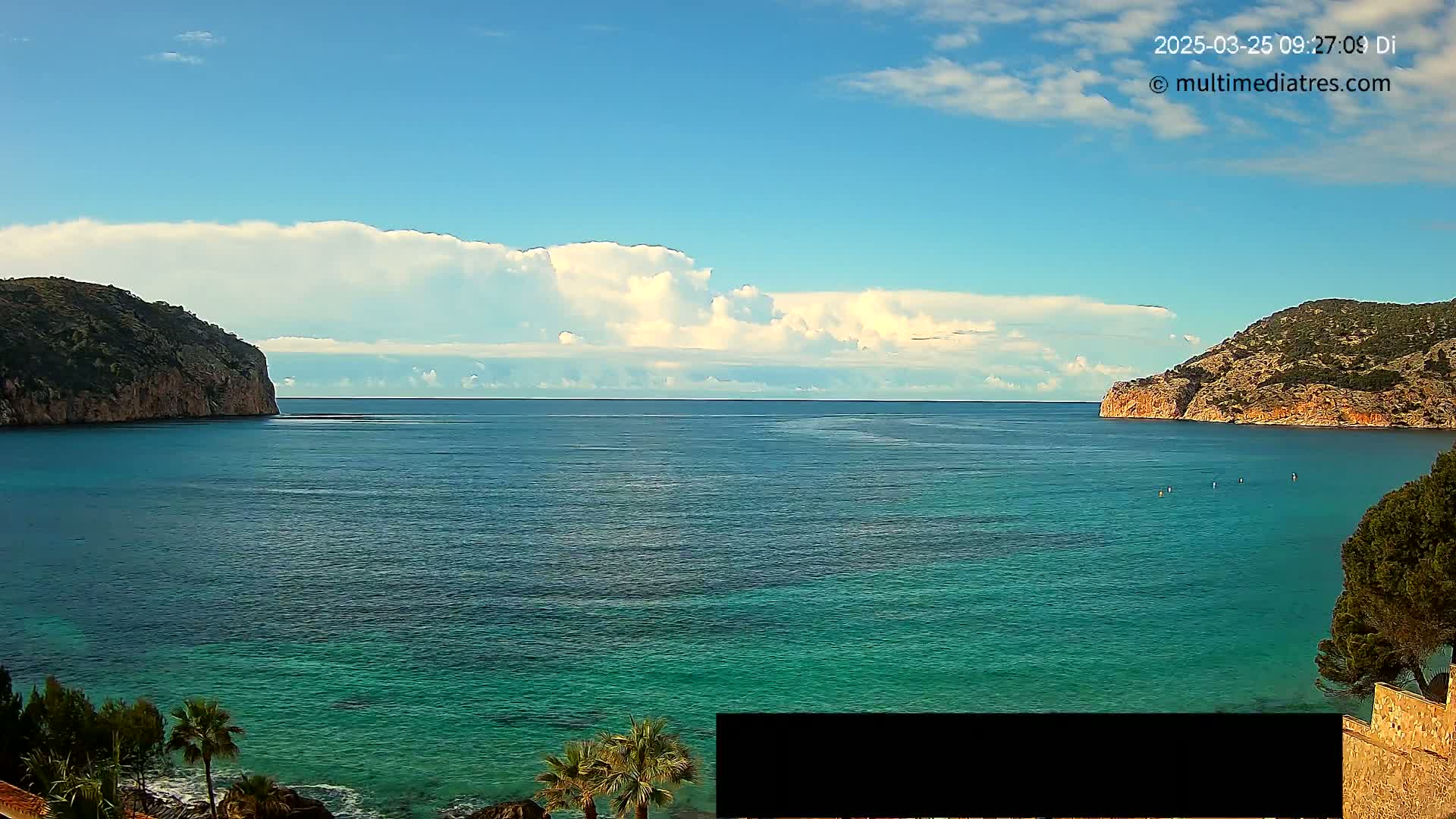 A calm, turquoise sea is nestled between two rocky hills under a partly cloudy sky.