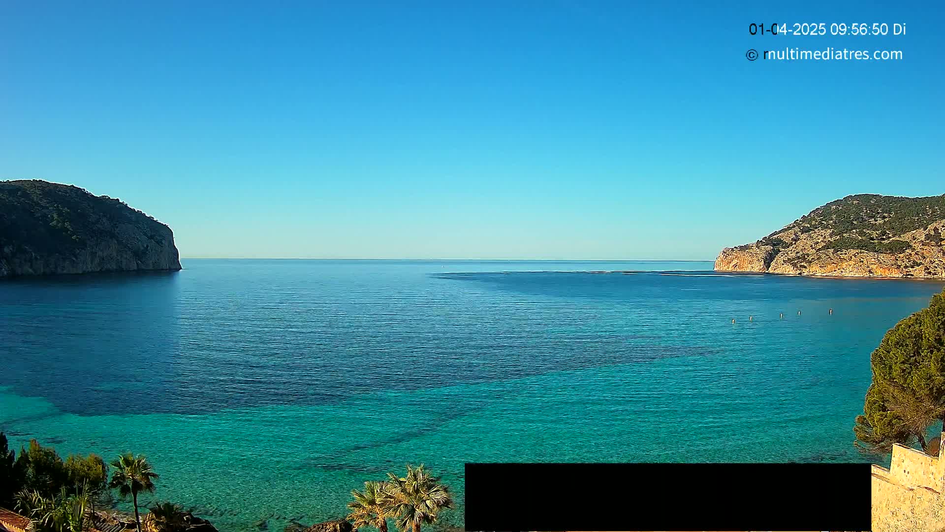 A clear blue sky overlooks a calm, turquoise ocean bay nestled between two rocky hillsides.