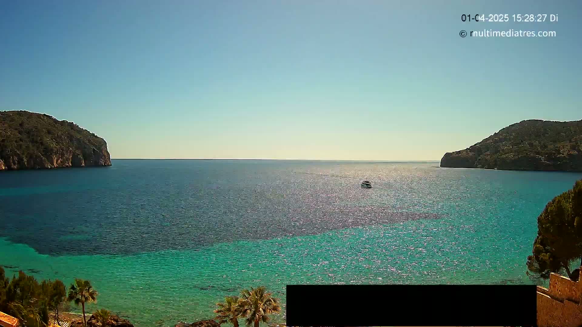 A calm, clear day shows a body of vibrant turquoise water nestled between two rocky hills, with a small boat visible in the distance.