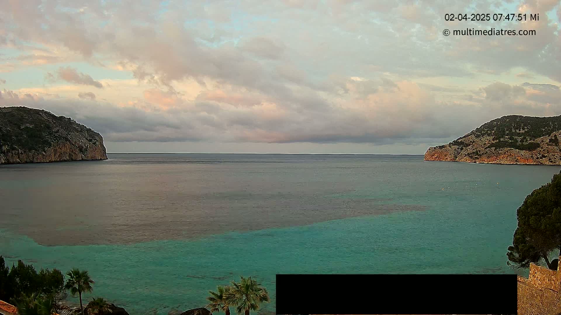 A calm bay with teal water is nestled between two rocky hills under a partly cloudy sky.