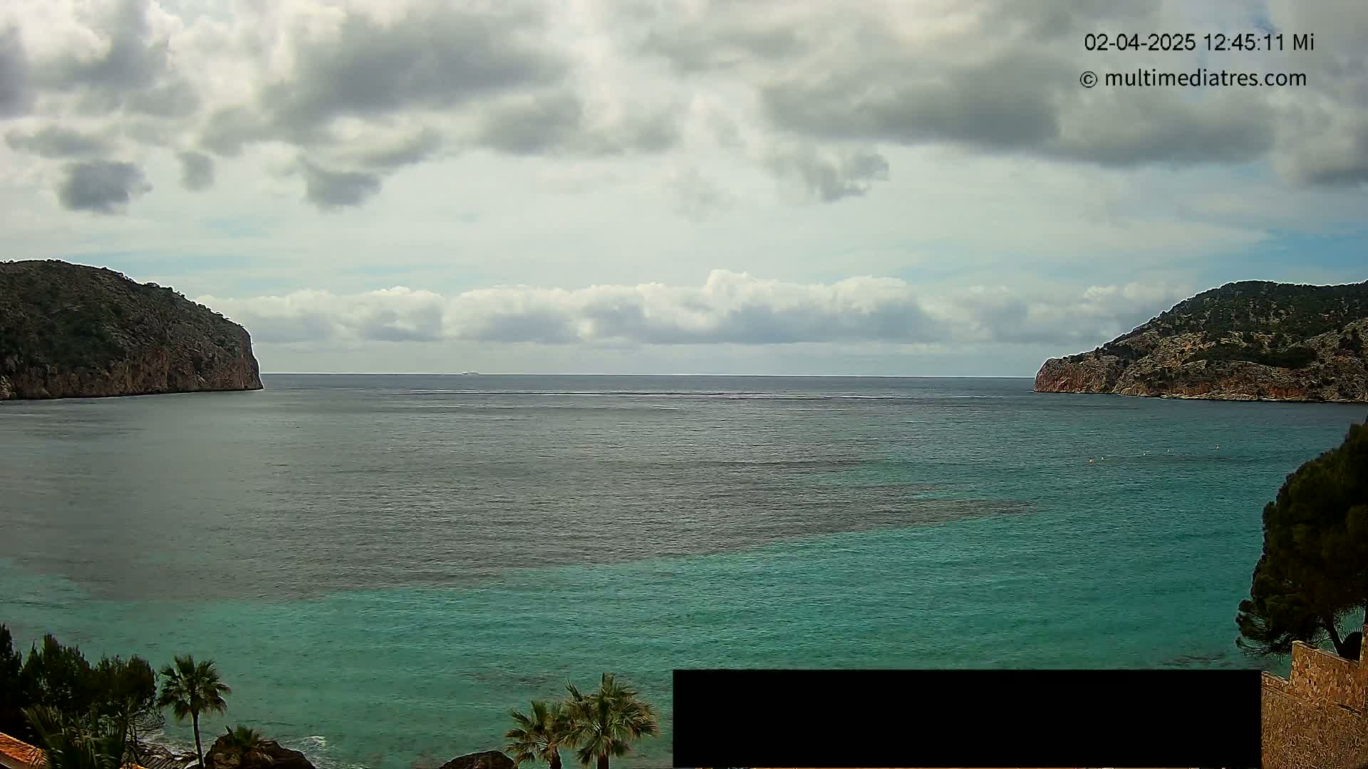 A partly cloudy day reveals a calm bay of teal water between two rocky hillsides, with some vegetation visible along the shoreline.