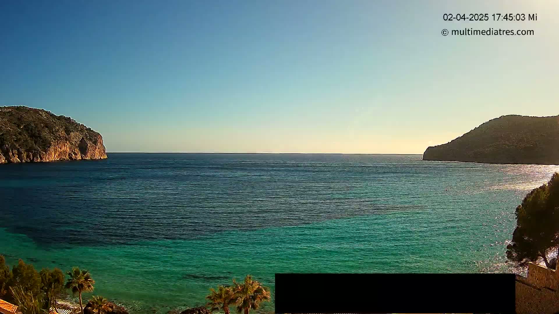 A calm, clear day reveals a picturesque cove with turquoise water nestled between two rocky hillsides, partially obscured by vegetation.