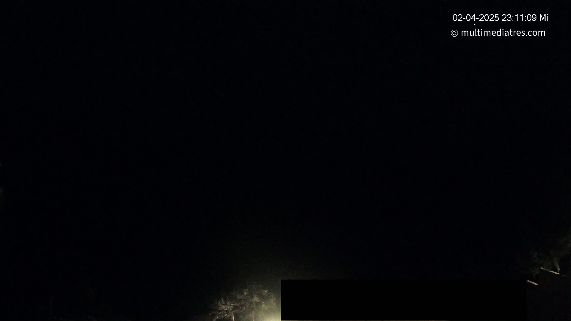 A nighttime, low-light image shows a dark sky and the faint illumination of some trees and a road in the lower portion of the frame.