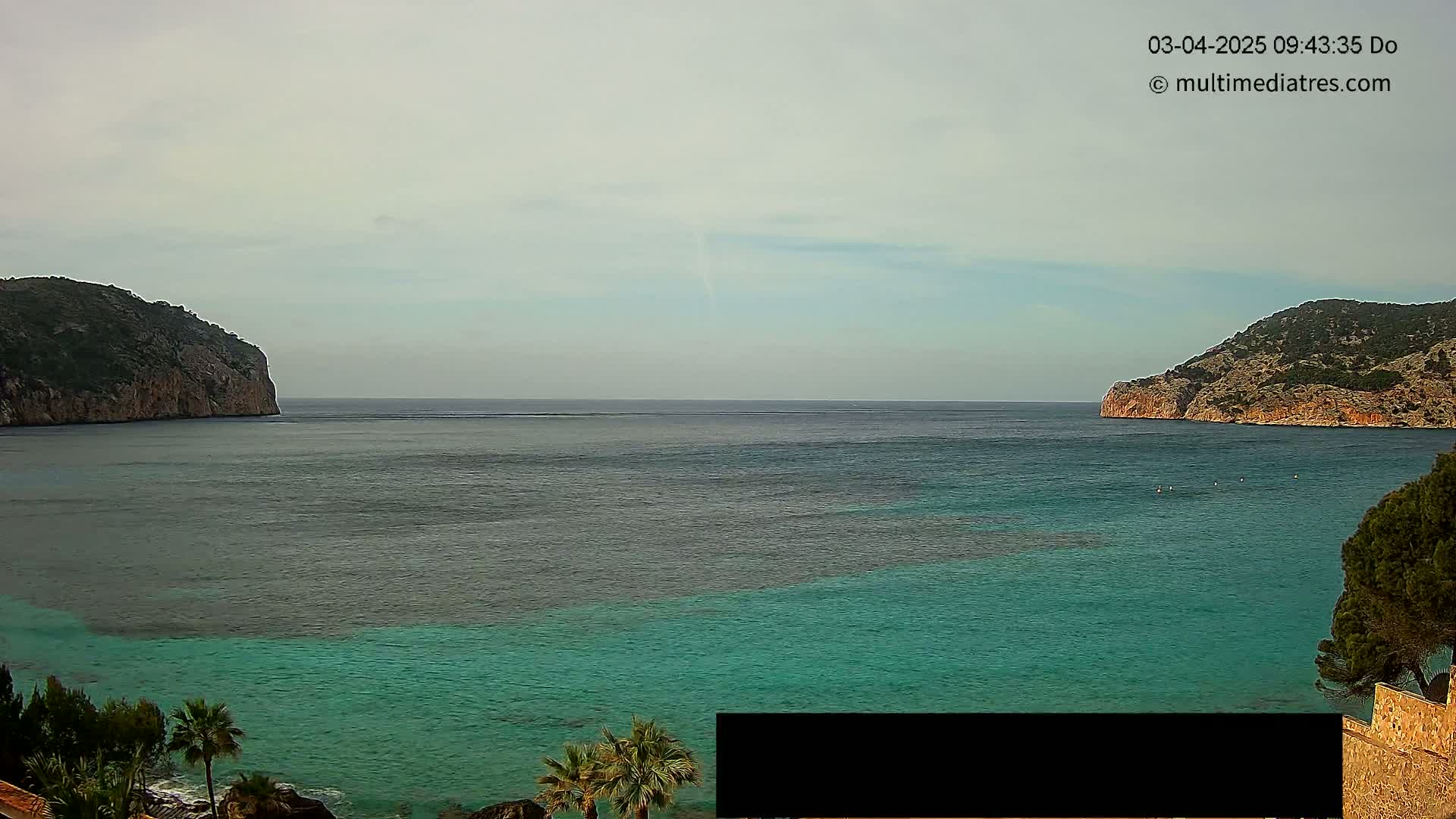 A calm, overcast day reveals a tranquil bay of teal water nestled between two rocky, green hills.