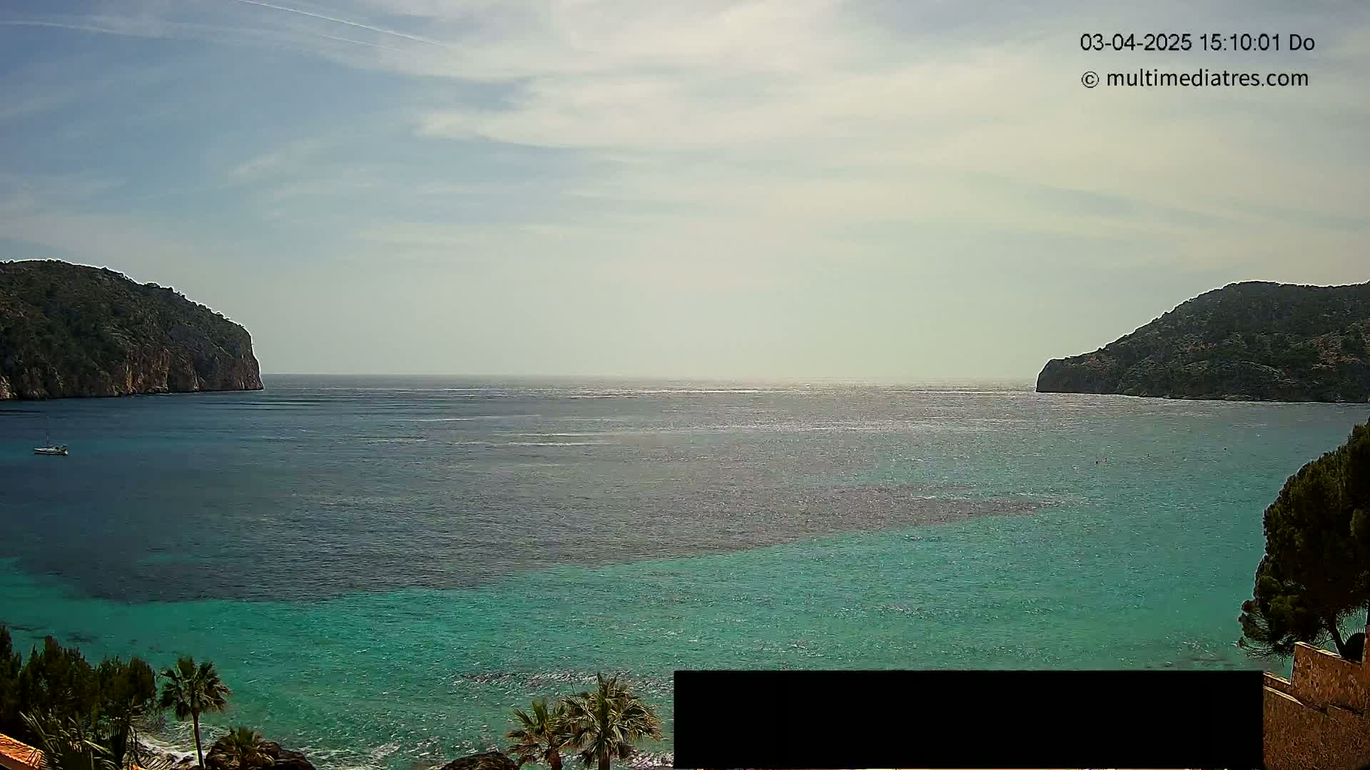 A tranquil bay of teal water is nestled between two rocky hills under a partly cloudy sky.