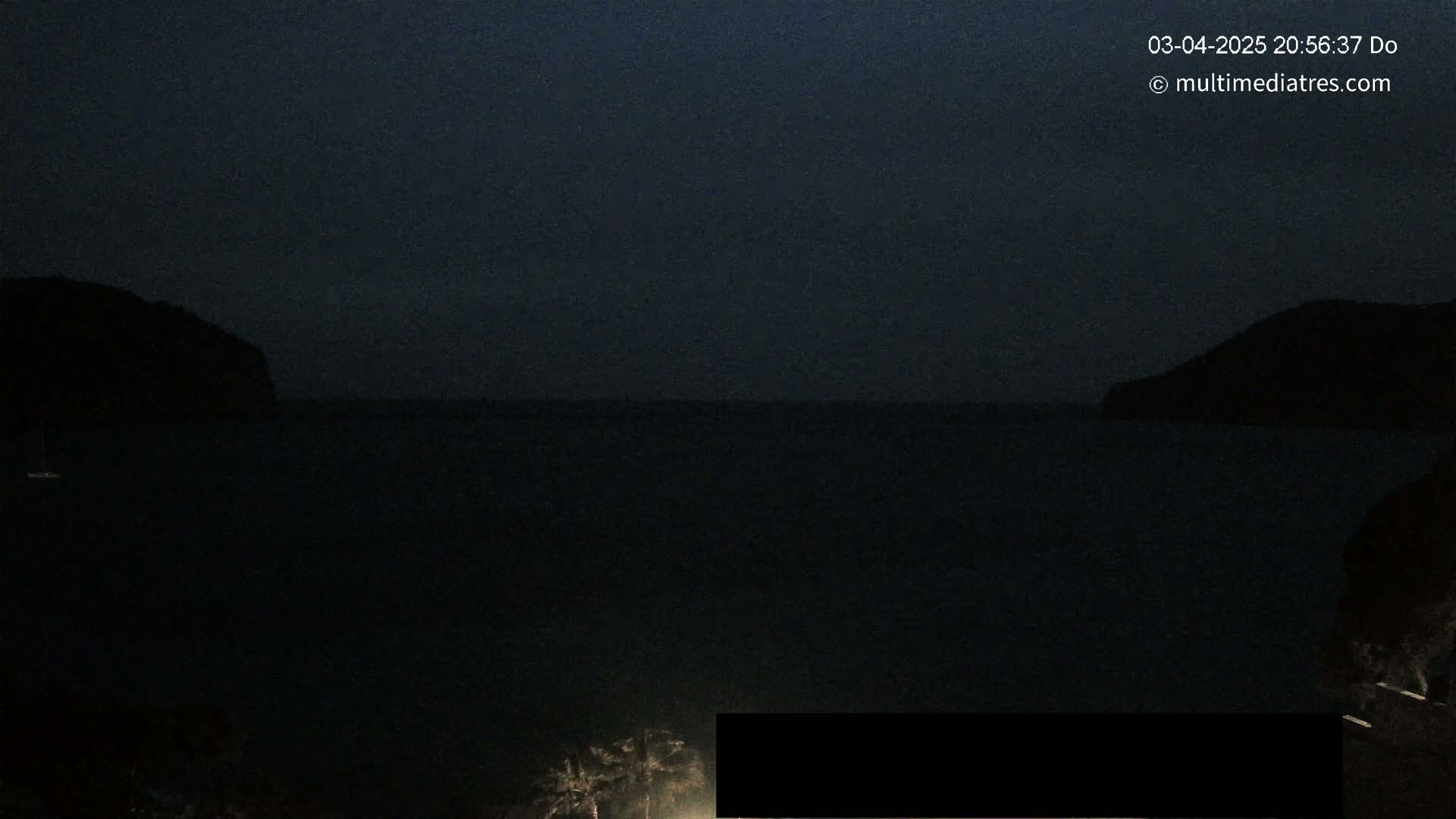 A dark, nighttime scene shows a body of water between two silhouetted hills under a clear, dark sky.