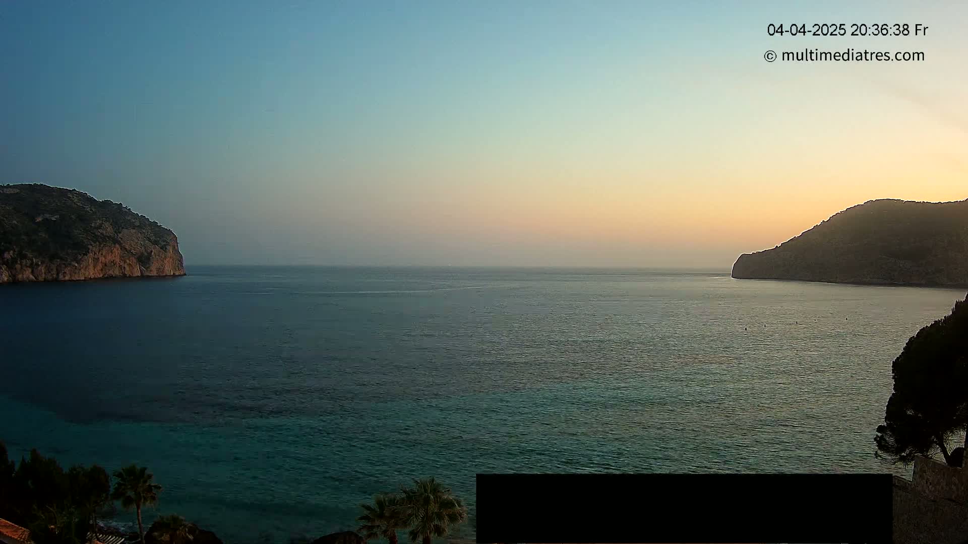 A calm sea at sunset is nestled between two dark, rocky hills under a clear, dusky sky.