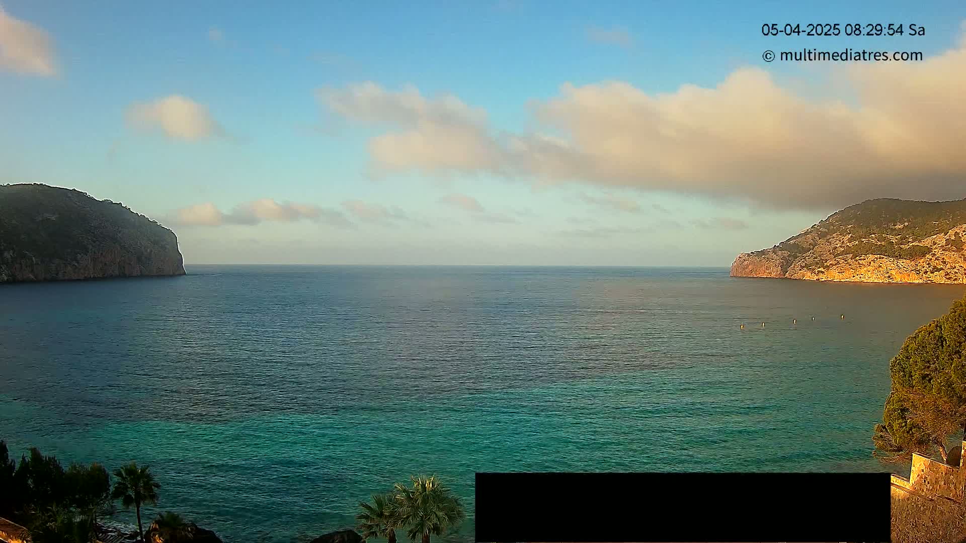 A partly cloudy morning scene shows a calm, teal ocean between two rocky hillsides with some vegetation.