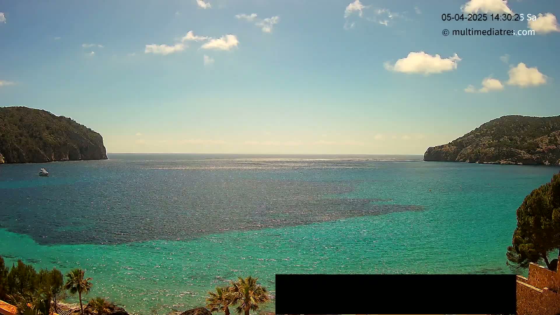 A partly cloudy sunny day reveals a tranquil cove of teal water between two rocky hills, with a single boat visible in the distance.