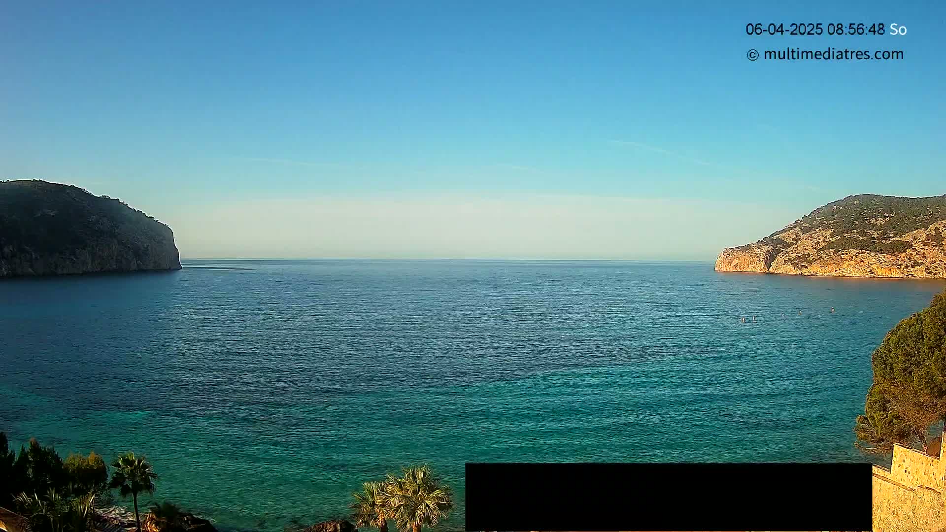 A calm, clear day reveals a picturesque coastal scene with teal water separating two rocky hills, where several people are paddleboarding in the distance.