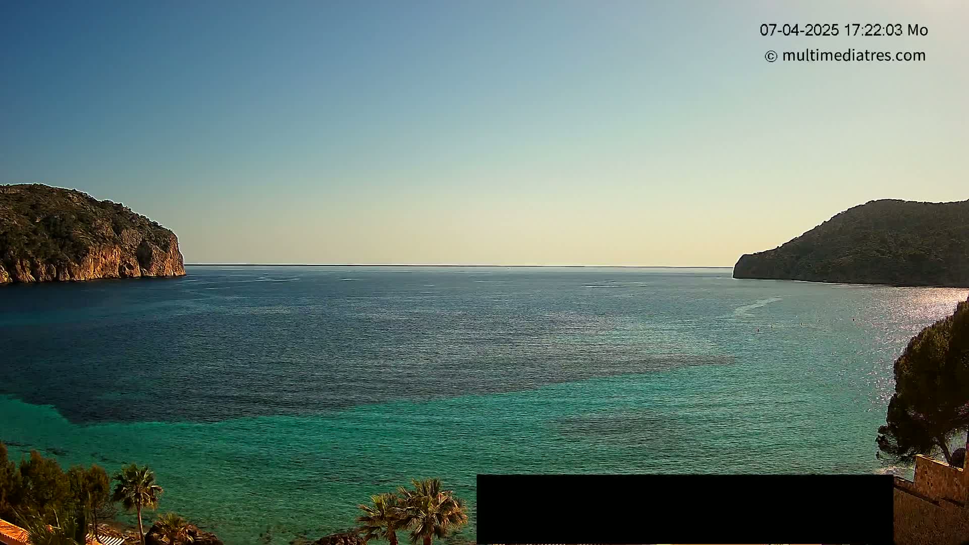 A tranquil coastal scene under a clear blue sky shows a calm, teal-colored sea between two dark green, rocky hills.