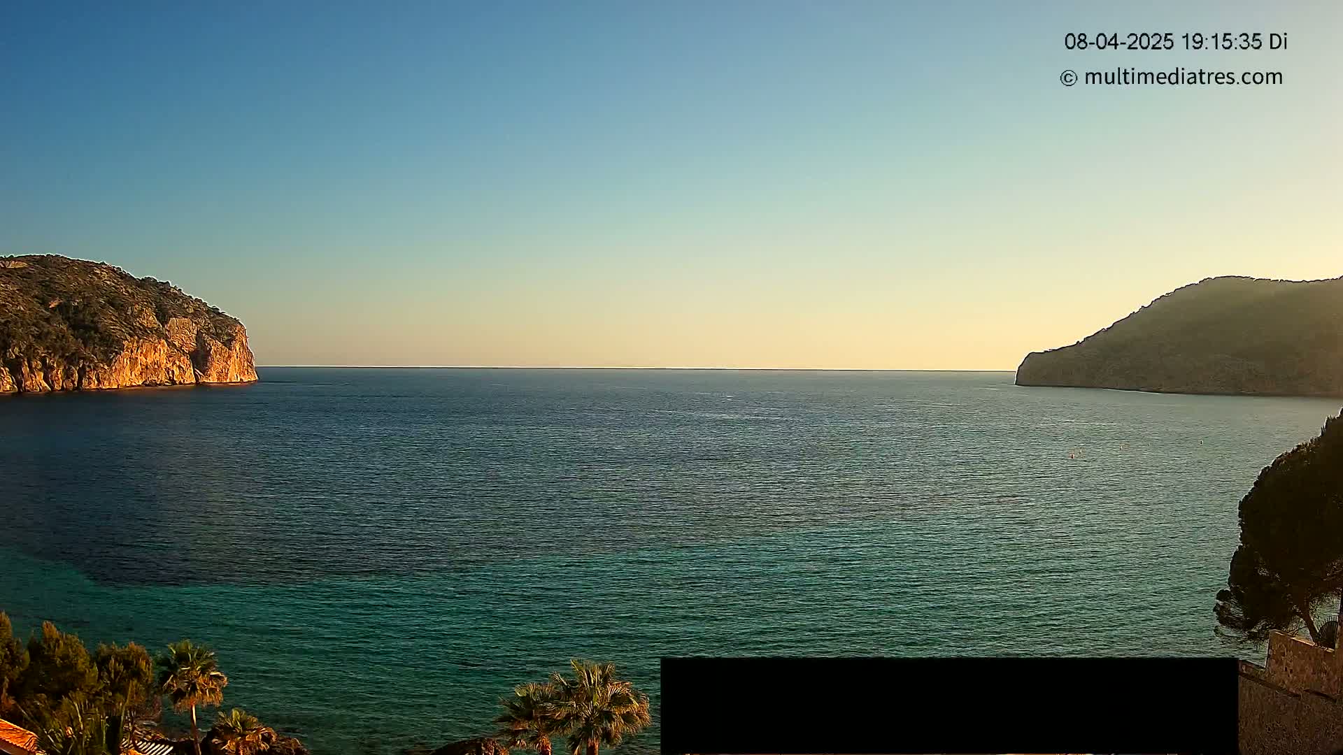 A calm, clear day shows a tranquil bay of dark teal water nestled between two rocky hillsides.